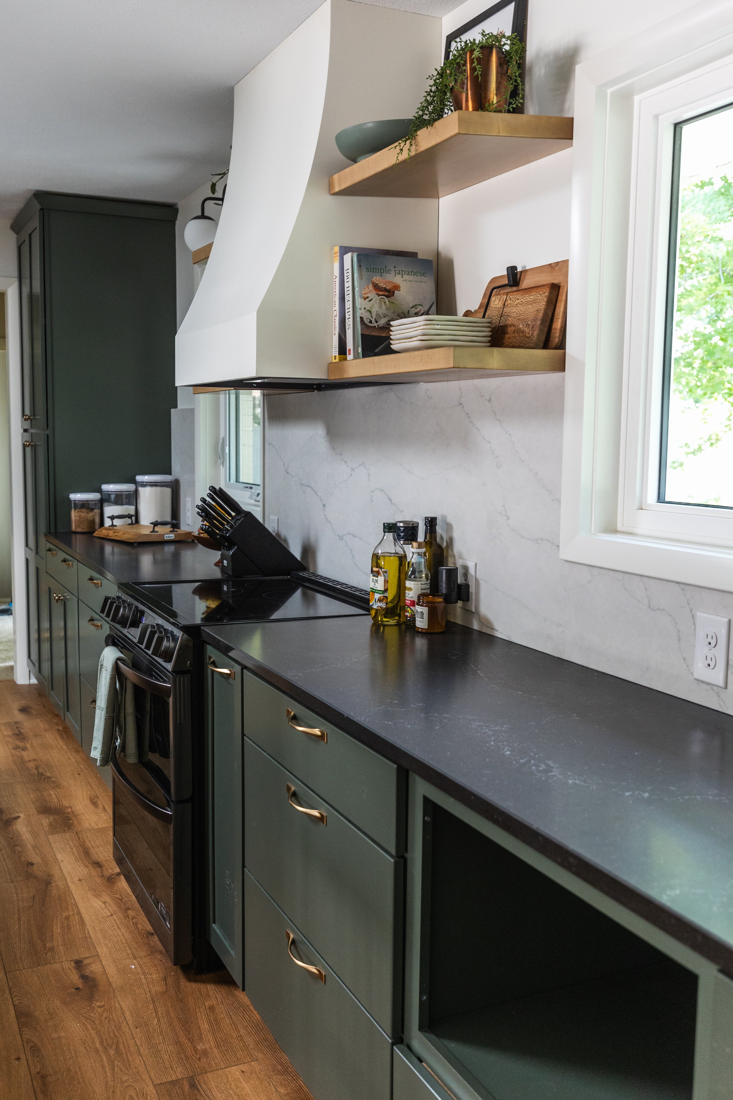 1962 Kitchen Remodel, dark green cabinets, white quartz island, wooden floors, brass hardware, wood cabinets, custom hood, floating shelves, dark countertops, open shelving, large windows, craftsmanship.