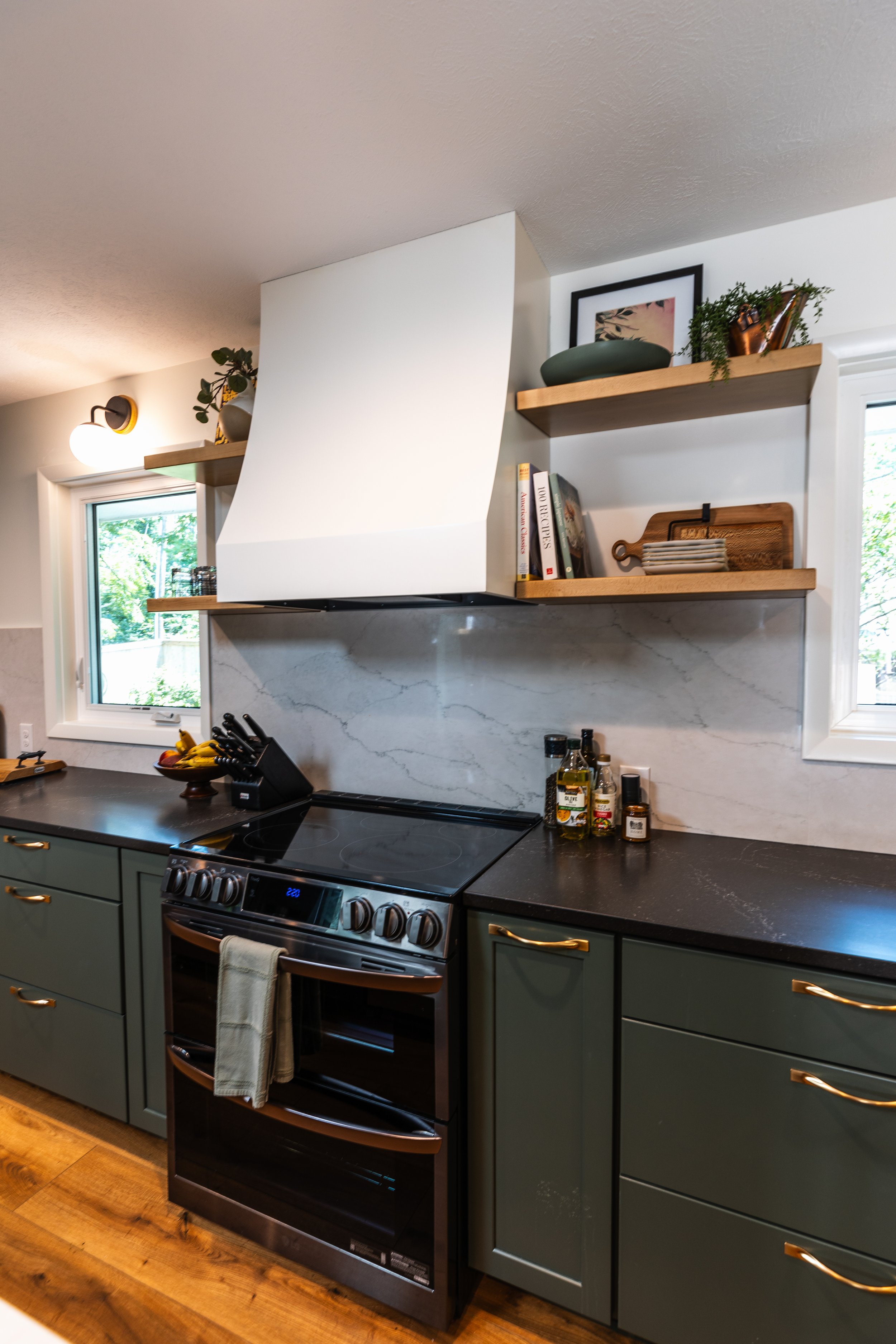1962 Kitchen Remodel, dark green cabinets, white quartz island, wooden floors, brass hardware, wood cabinets, custom hood, floating shelves, dark countertops, open shelving, large windows.
