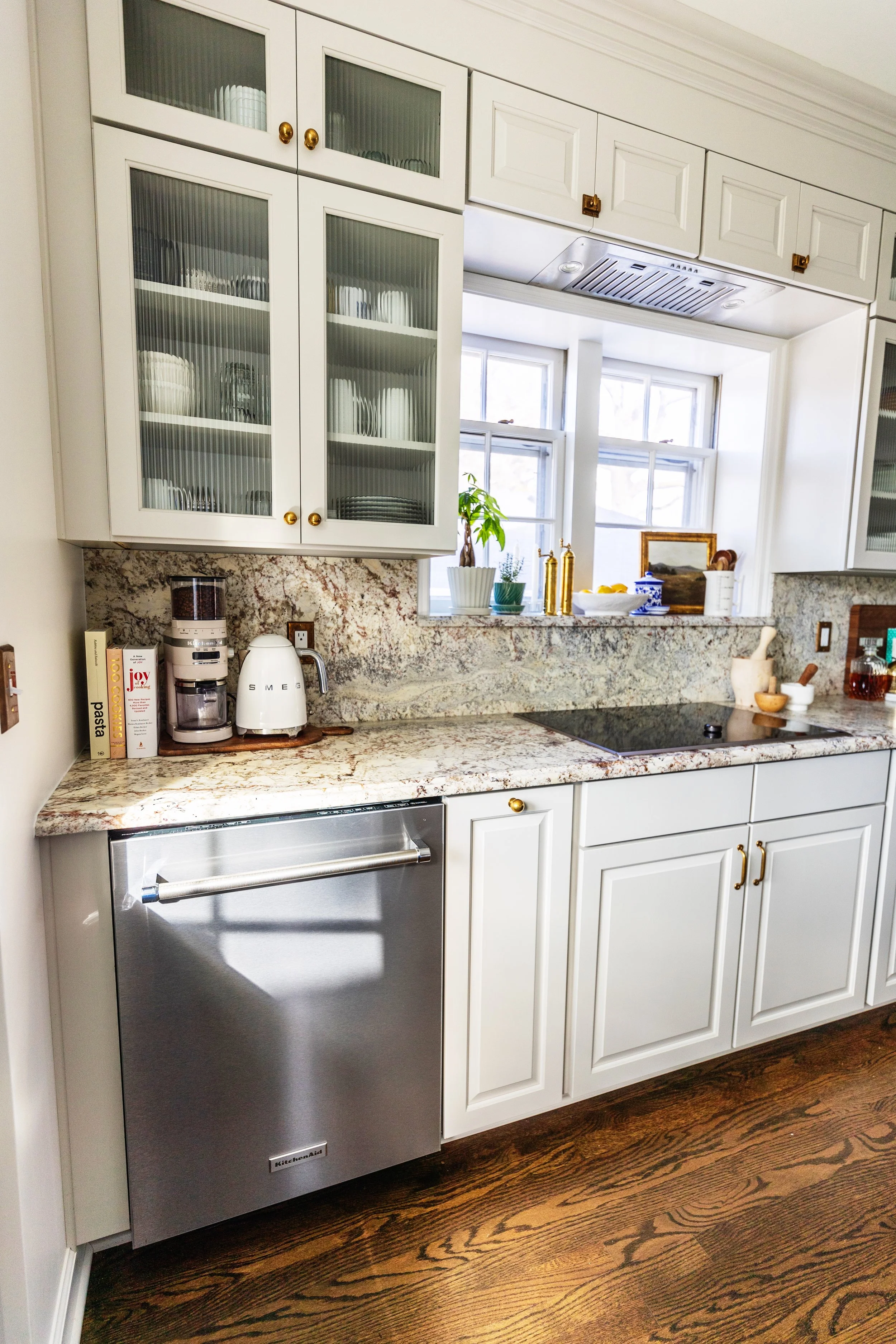 Kitchen with white cabinets, granite countertop, under-the-counter dishwasher, coffee machine, and window with plants on the windowsill.
