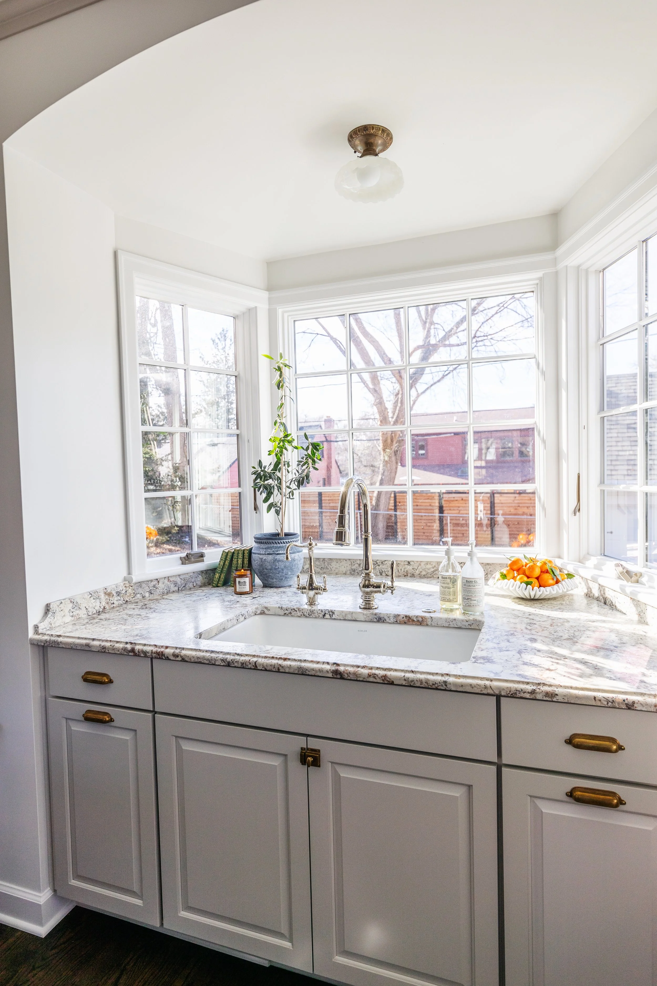 Kitchen window over a marble countertop with a potted plant, soap dispensers, and a bowl of oranges.