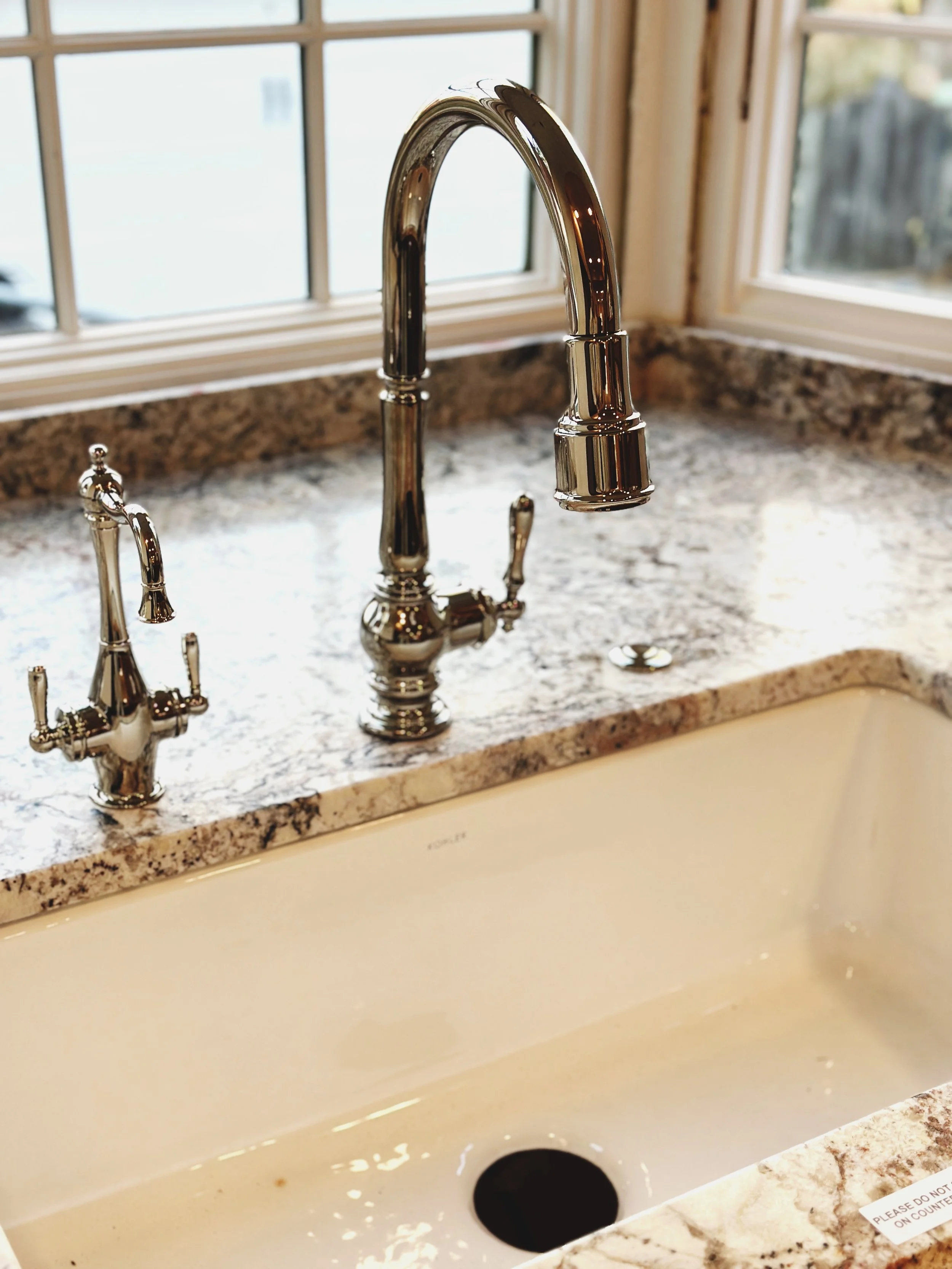 Kitchen sink with chrome faucet and soap dispenser, granite countertop, window in the background.
