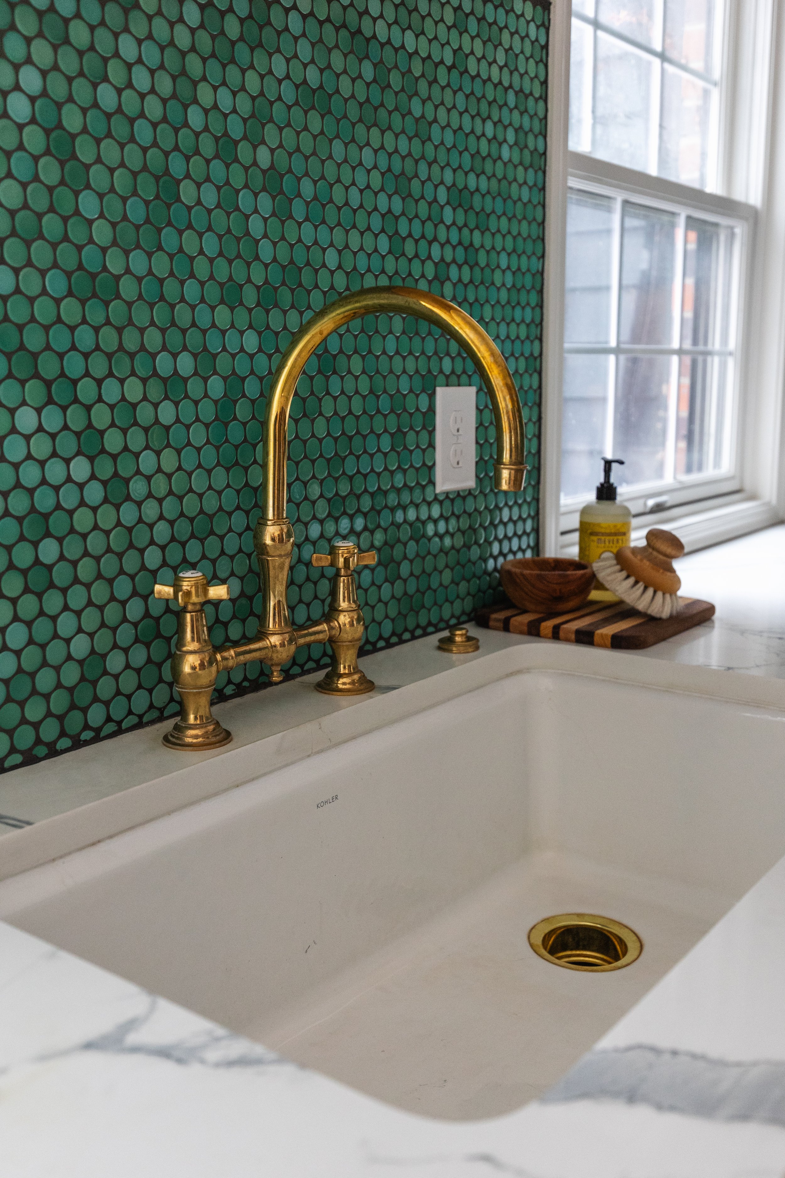 Close-up of a white kitchen sink with gold faucet and handles, green dotted tile backsplash, soap dispenser, wooden bowl, and cleaning brush on a black striped wooden tray next to a window.