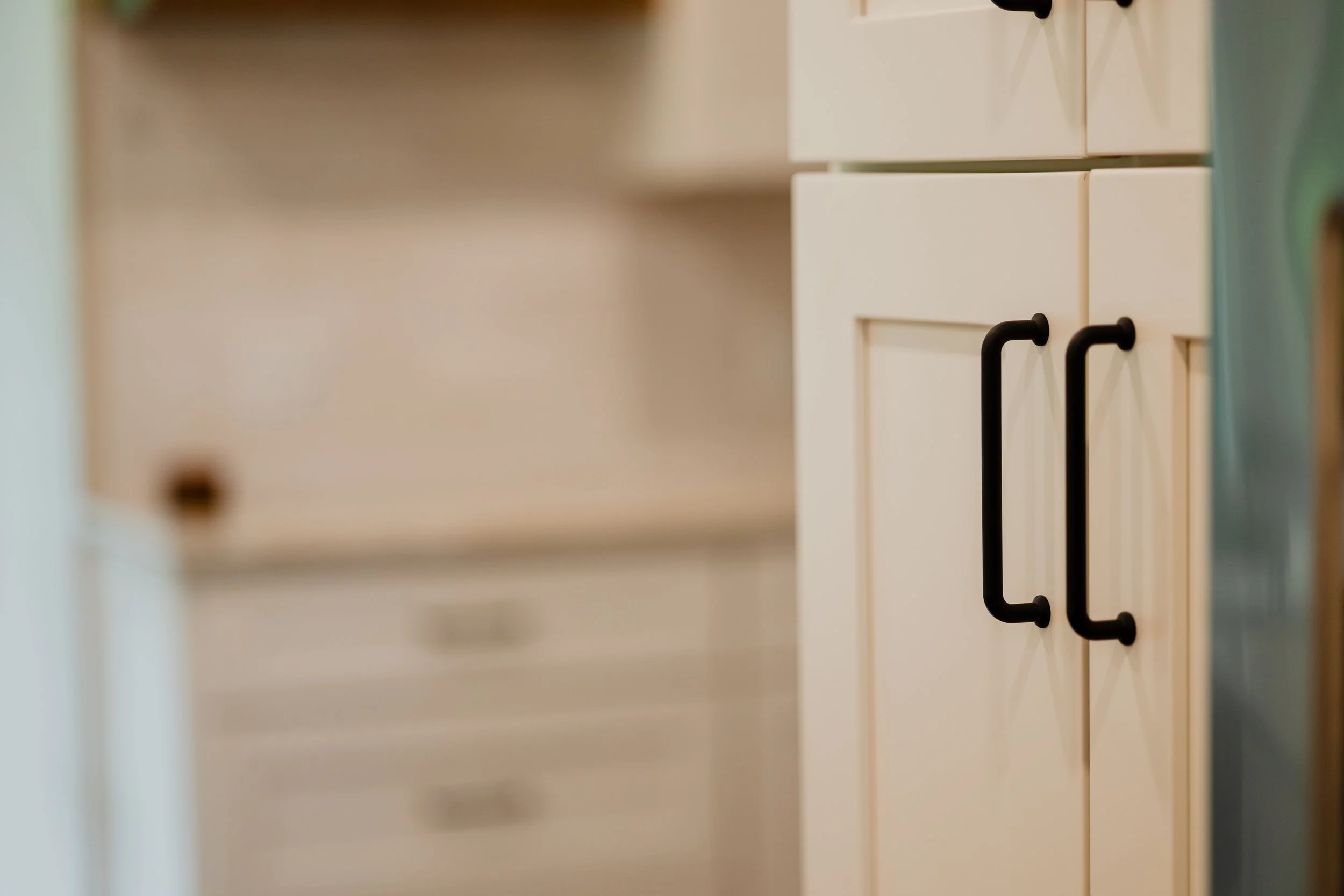 Close-up of white kitchen cabinet doors with black handles, blurred surrounding cabinets. 1935 Kitchen Remodel. Black Matte Hardware. White Cabinets.