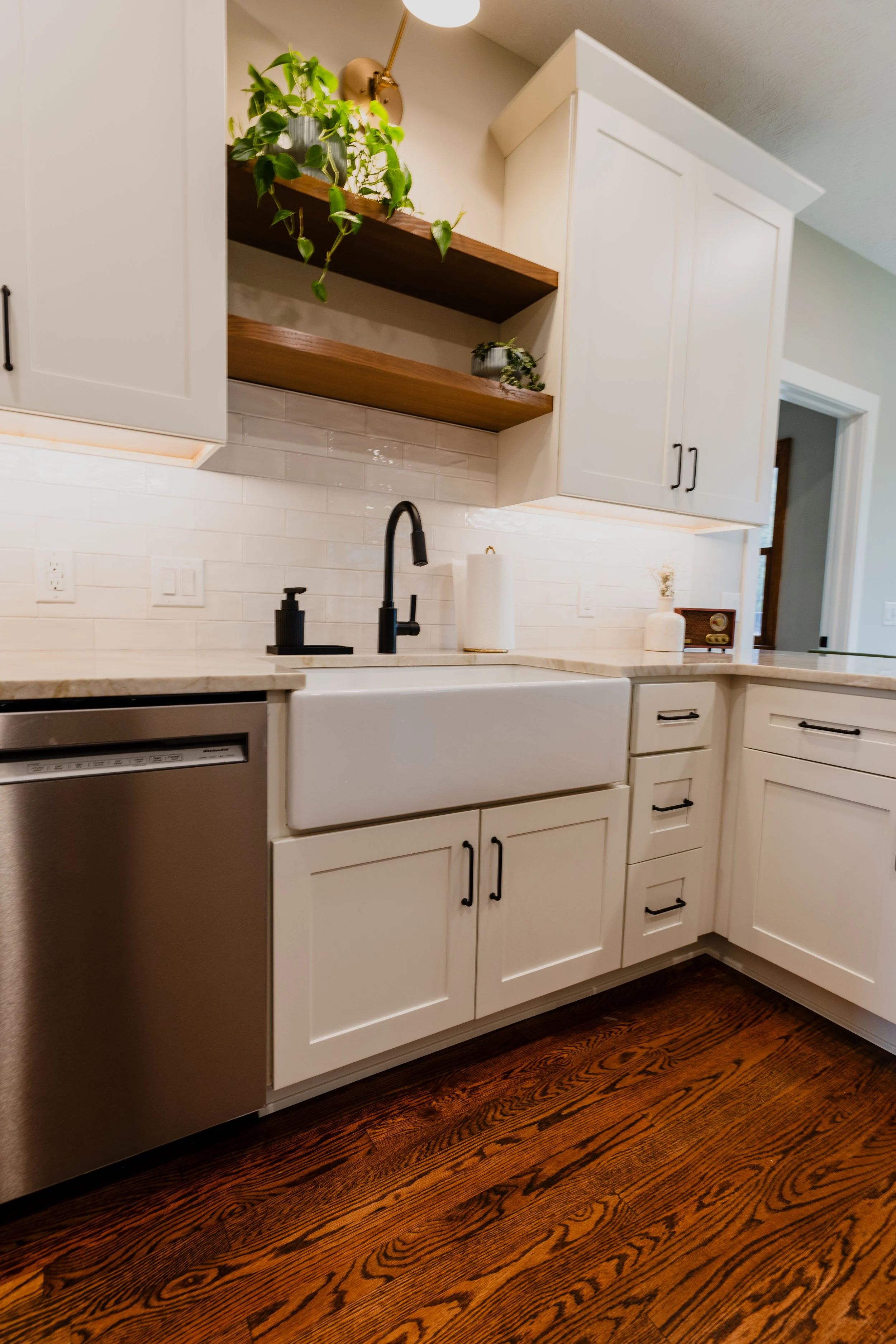 Kitchen with white cabinets, black faucet, farmhouse sink, wood open shelves with plants, white tile backsplash, stainless steel appliance, and hardwood floor.