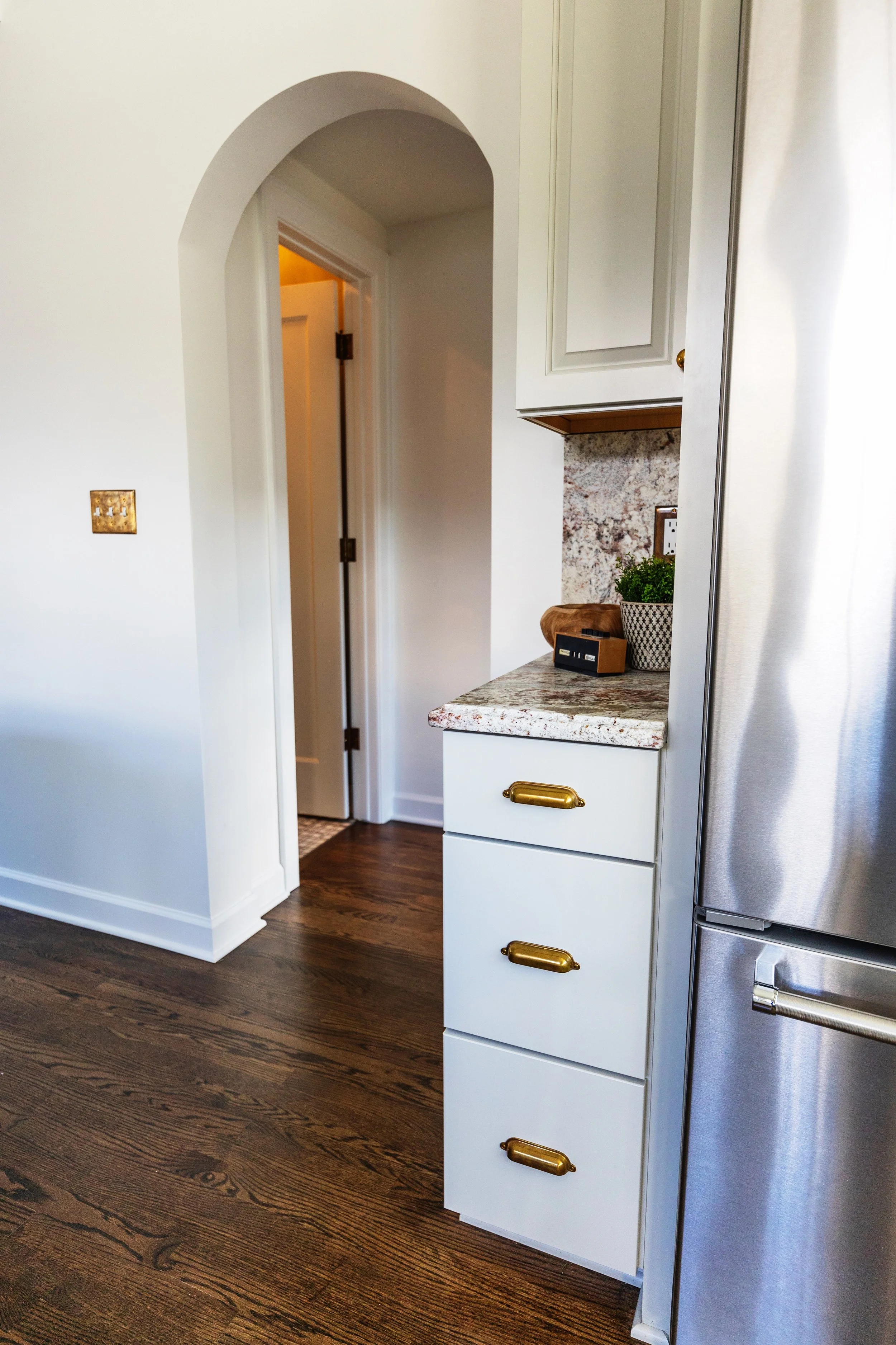 Kitchen with white cabinets and drawers with brass handles, dark hardwood flooring, granite countertop, and a partial view of a stainless steel refrigerator. An arched doorway leads to a room with a doorway illuminated by warm light.