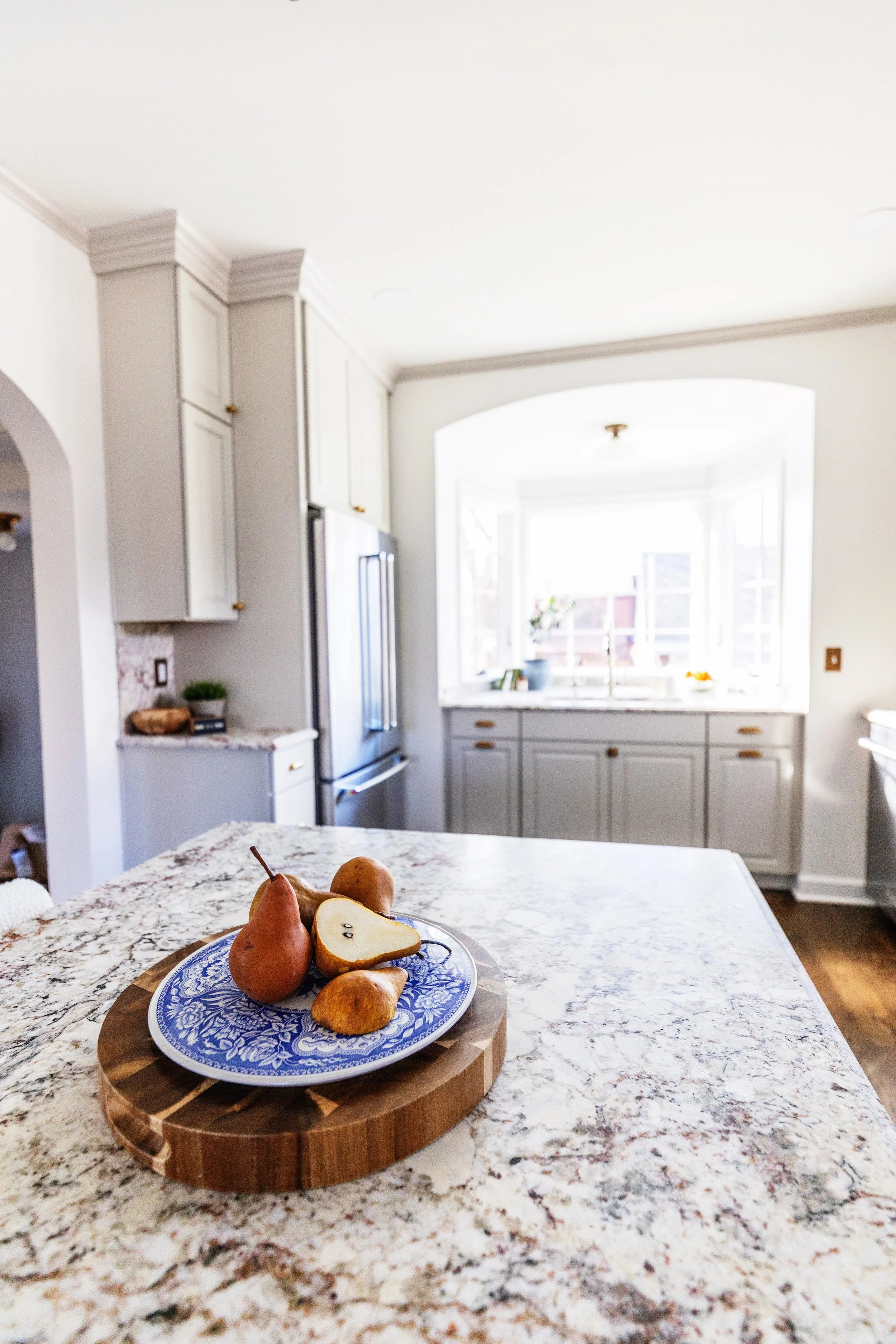 A kitchen with a marble countertop, white cabinets, and a large window with sunlight. A blue and white plate with pears and apples is on the island.