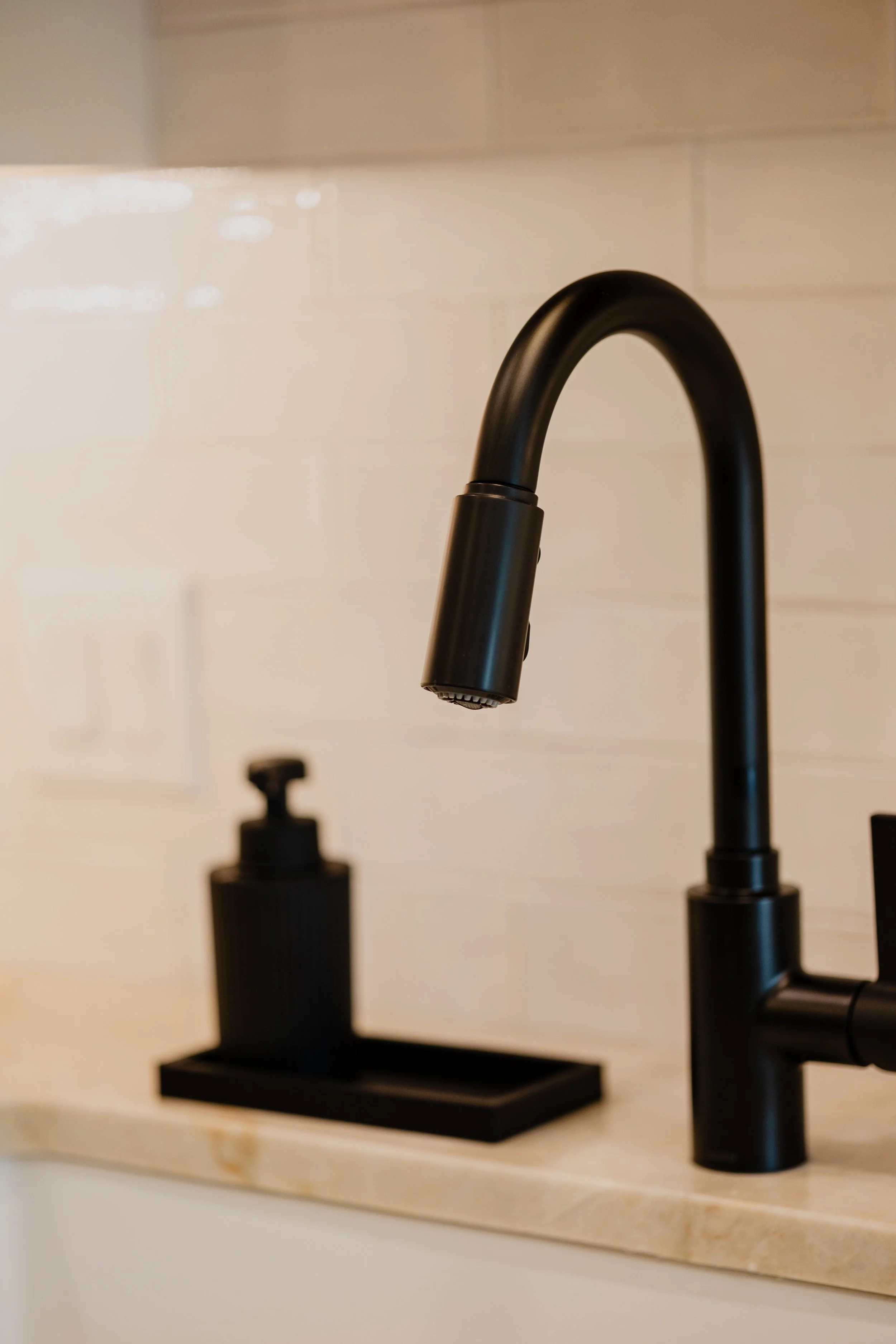 Close-up of a black kitchen faucet and a black soap dispenser on a beige countertop against a white tile wall. 1935 Kitchen Black Matte Faucet. White Subway Tile.