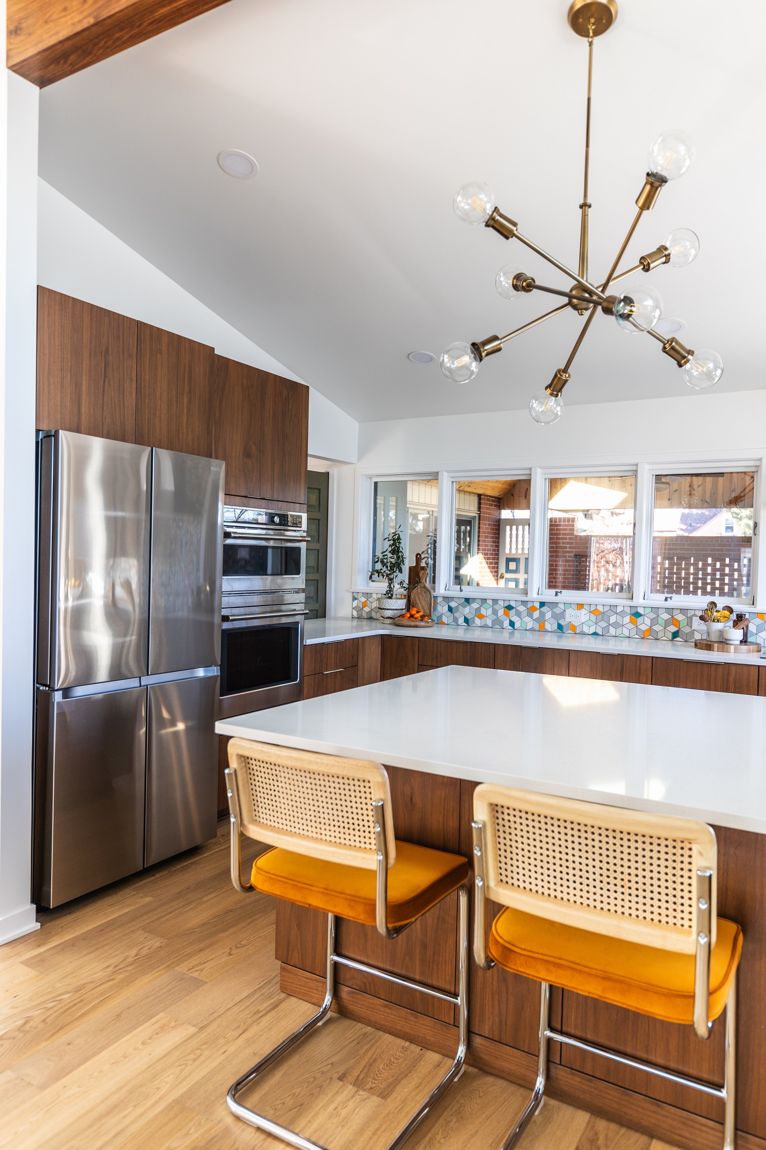Modern kitchen with a wooden accent wall, stainless steel refrigerator, built-in oven, white countertop, colorful tile backsplash, and two vintage-style bar stools with mustard cushions.