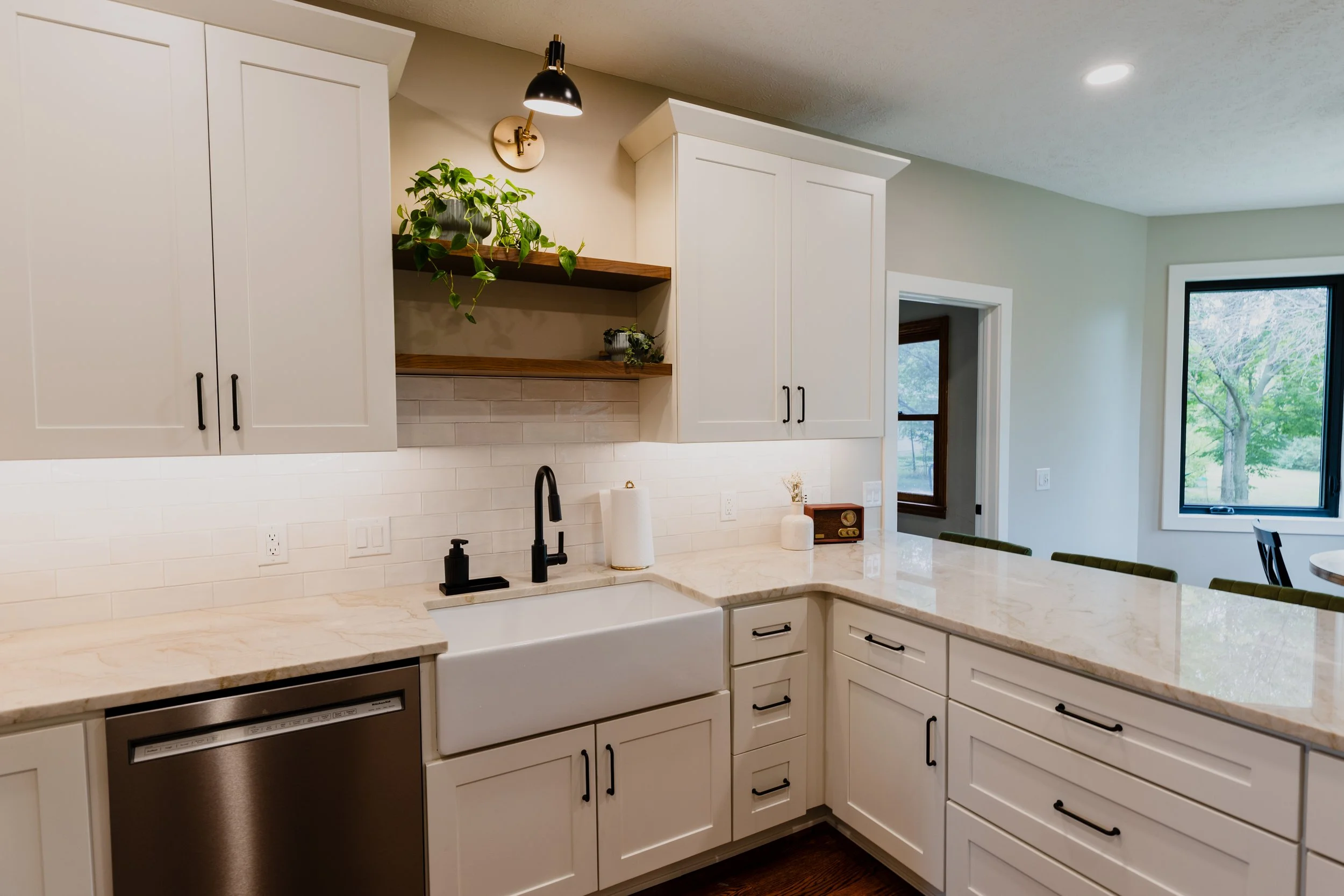 Modern kitchen with white cabinets, black handles, marble countertops, a farmhouse sink with black fixtures, a backsplash with white subway tiles, open shelves with plants, and a window showing greenery outside.
