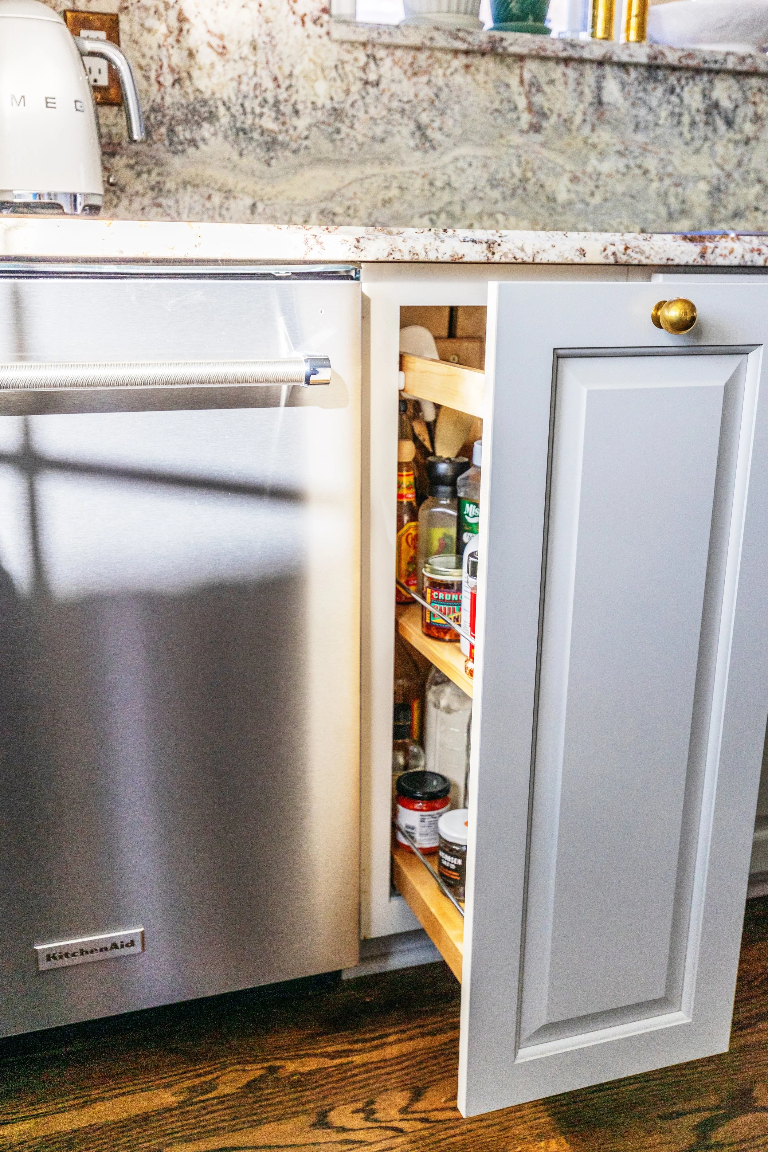 Open kitchen cabinet with spice jars and containers, next to a stainless steel appliance and granite countertop.