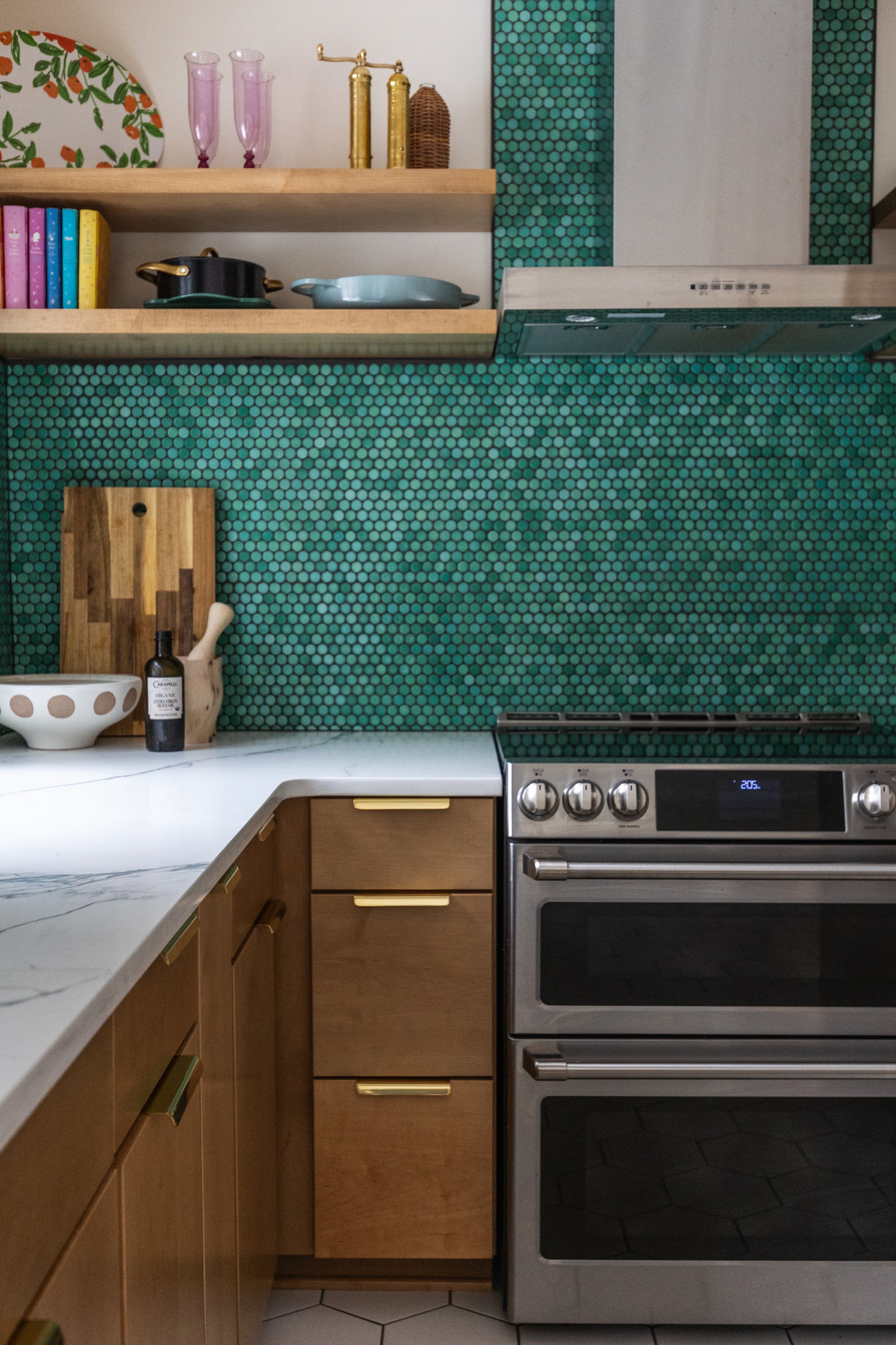 Kitchen with wood cabinets, a marble countertop, green round tile backsplash, open wooden shelves with dishes and glasses, and a stainless steel oven.