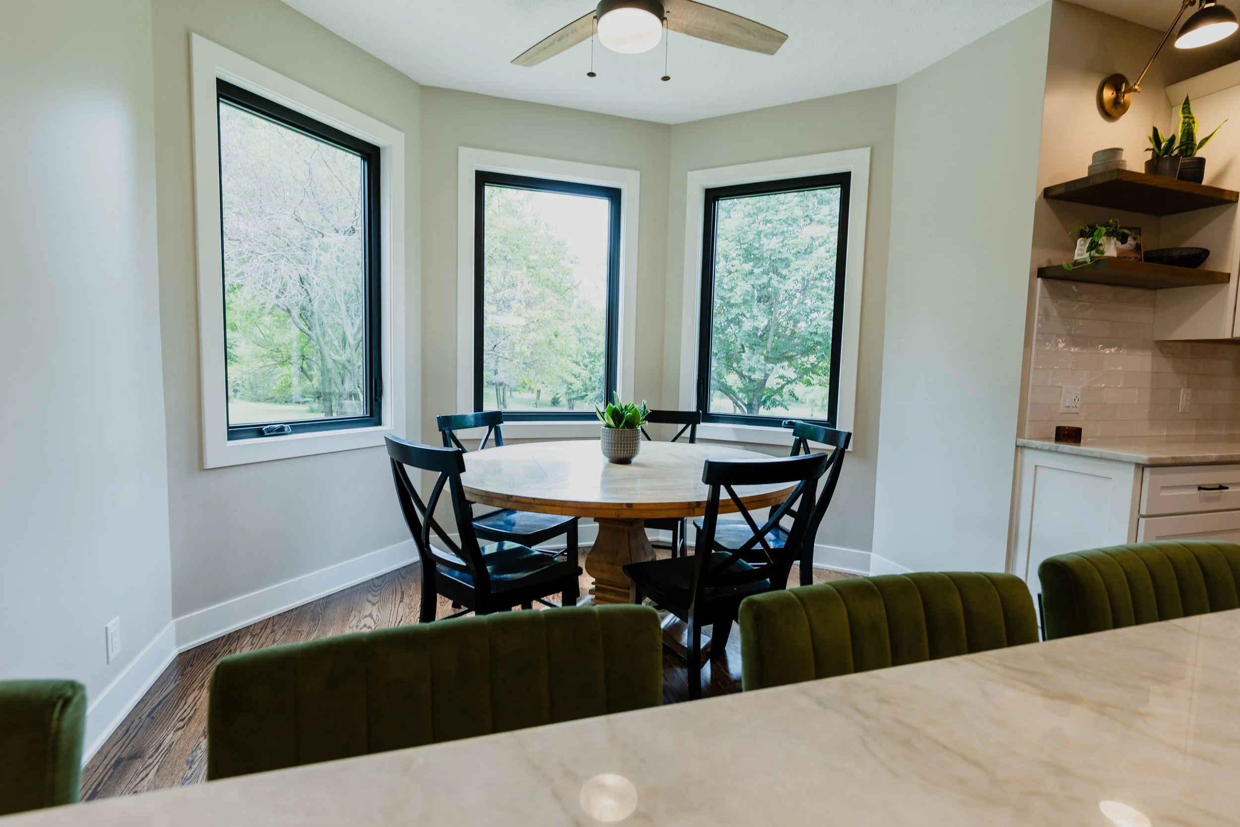 A kitchen nook with a round wooden table surrounded by five black chairs positioned near three large windows with trees outside. The room has light-colored walls, a white ceiling with a ceiling fan, and hardwood floors. part of a kitchen with green c