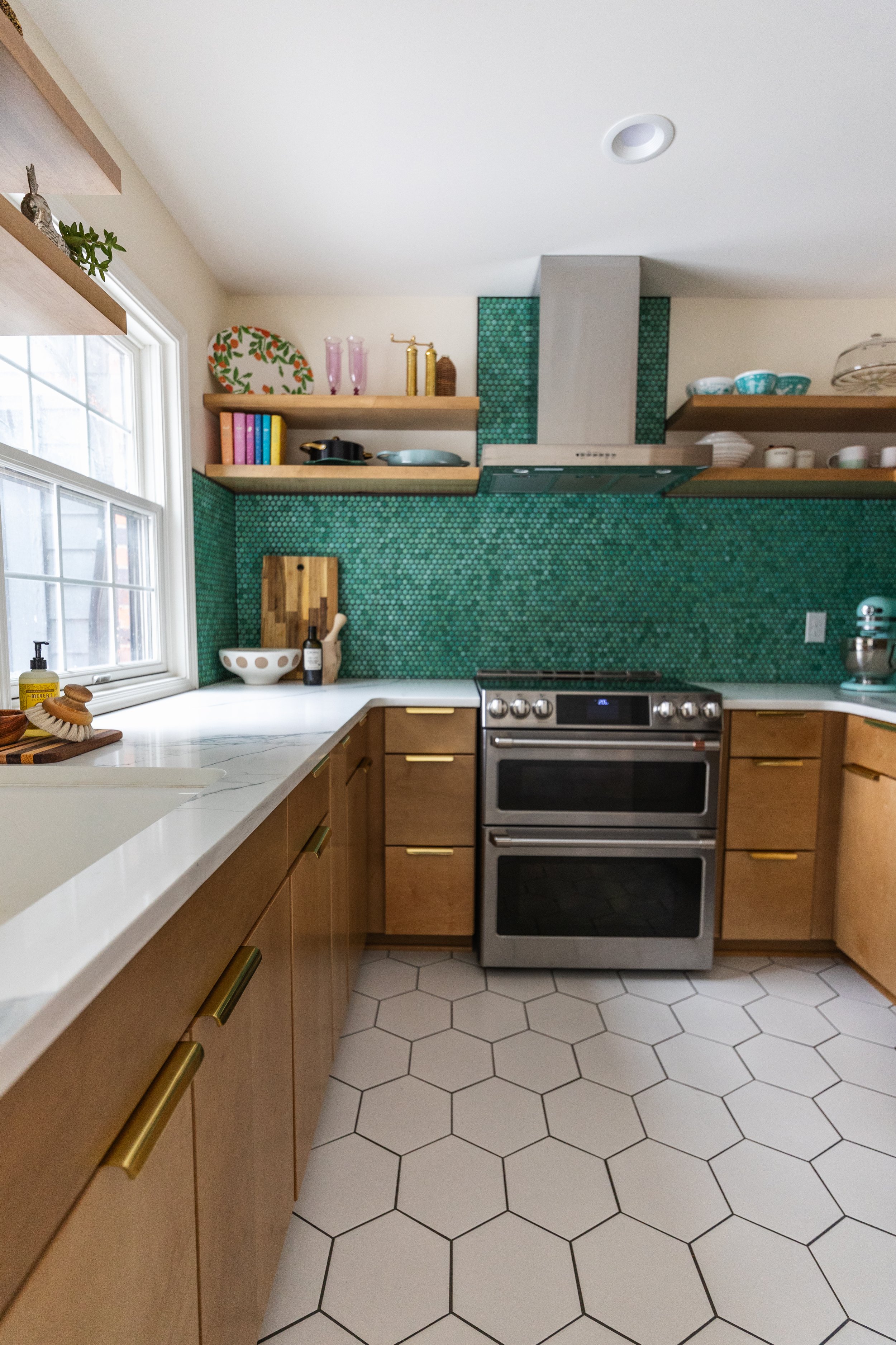 Modern kitchen with wooden cabinets, hexagonal white floor tiles, green mosaic tile backsplash, stainless steel stove, open shelves with dishes and decor, window with natural light.