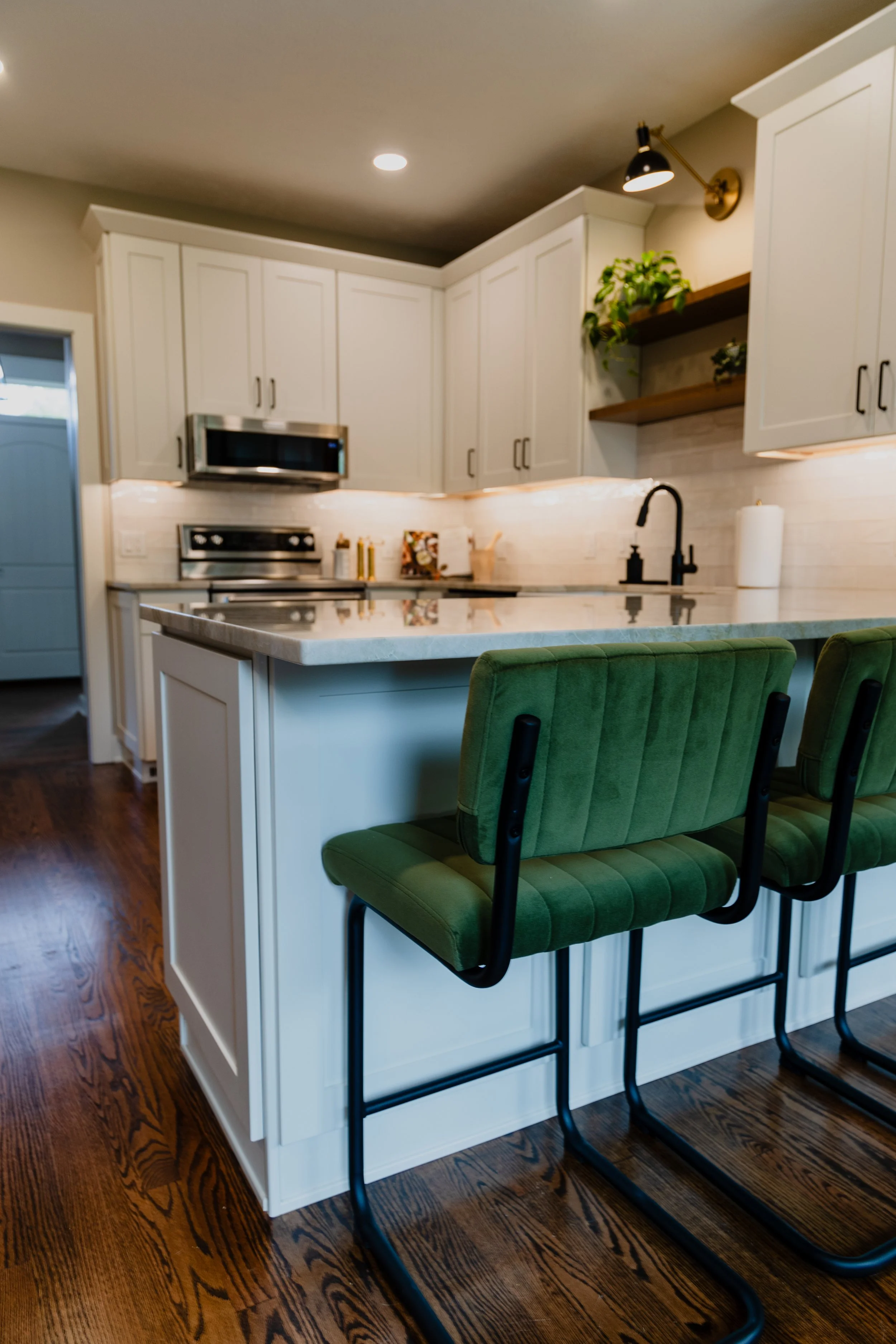 Modern kitchen with white cabinets, a marble countertop on a kitchen island, green upholstered barstools, and hardwood flooring. 1935 Kitchen Remodel