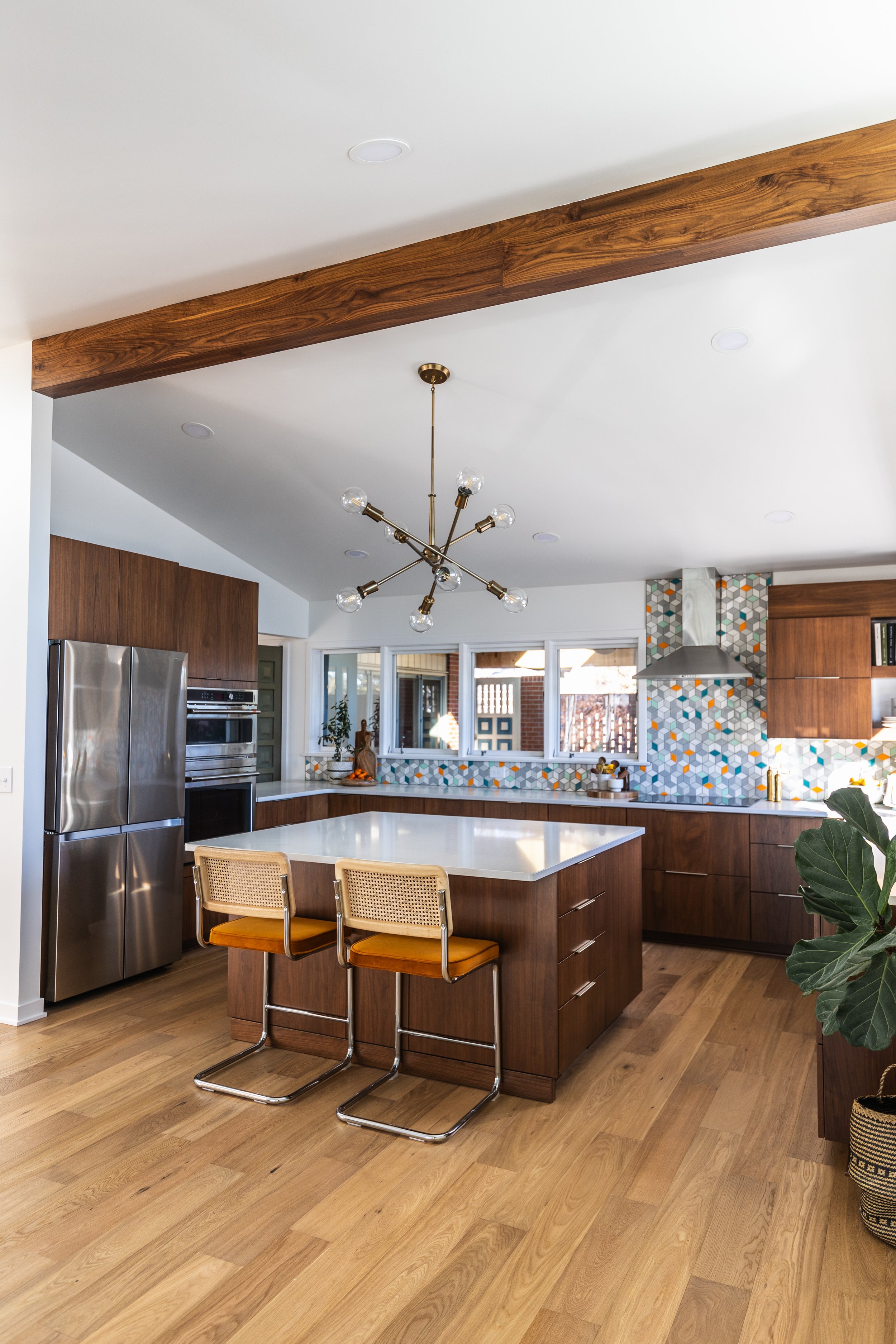 Modern kitchen with wooden cabinetry, a large kitchen island with a white countertop, two rattan barstools with orange cushions, stainless steel refrigerator, geometric patterned backsplash, and a contemporary brass chandelier.