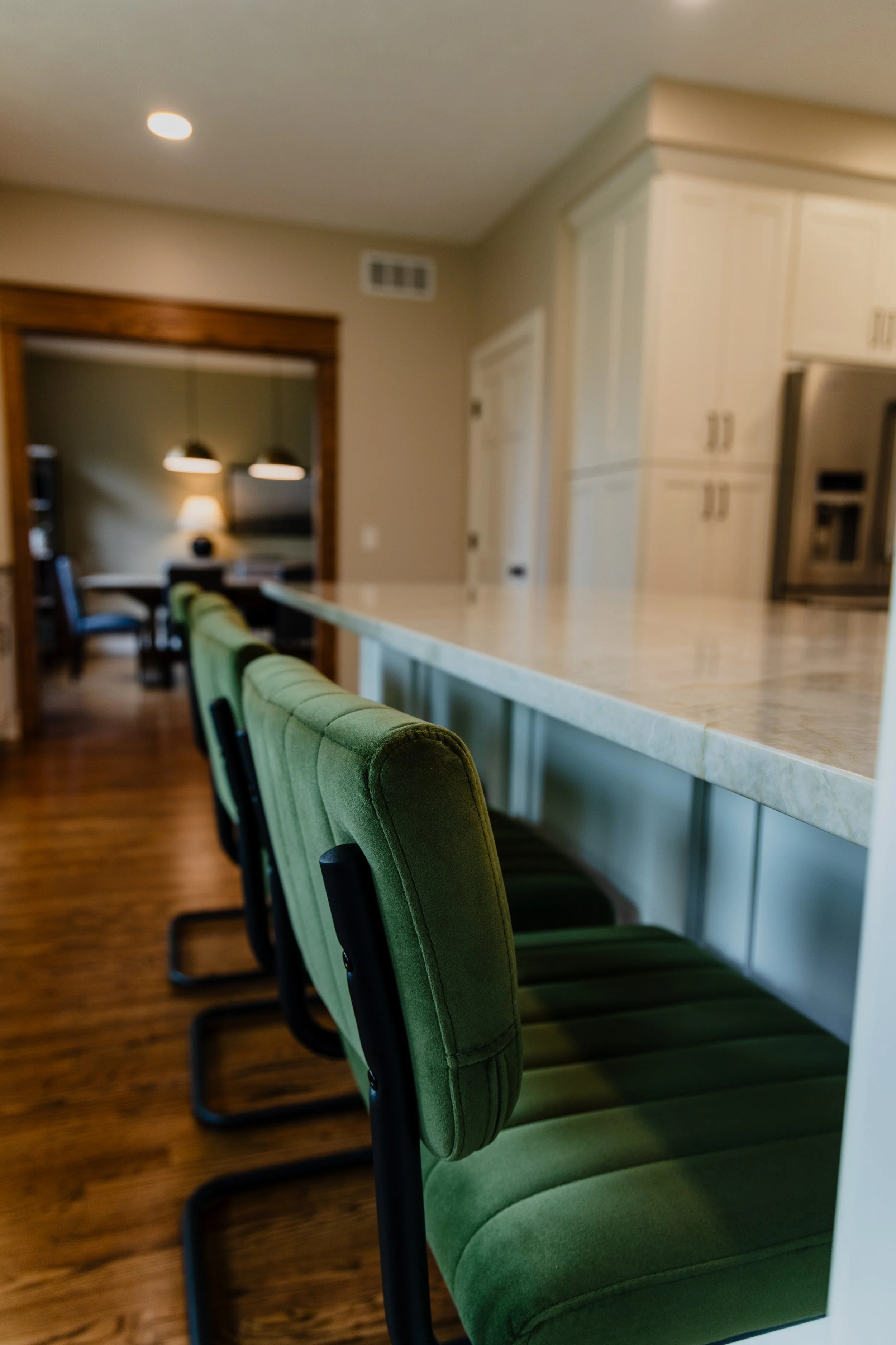 Green upholstered chairs positioned along a kitchen counter with a marble top in a modern home interior. 1935 Kitchen Remodel. Kitchen Island. Eat at bar.