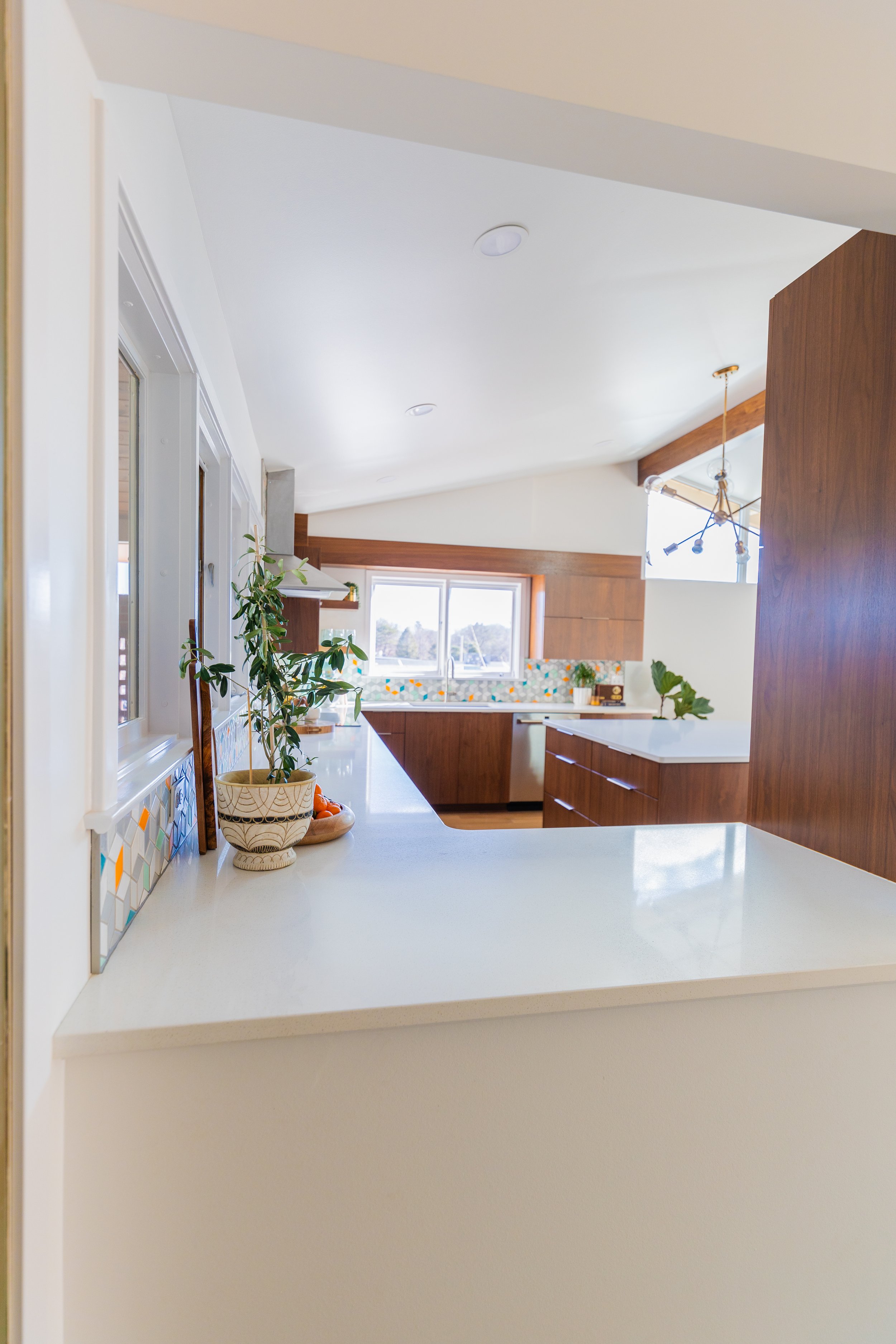 Modern kitchen with wooden cabinets, white countertops, colorful tile backsplash, large window, and a potted plant on the counter.