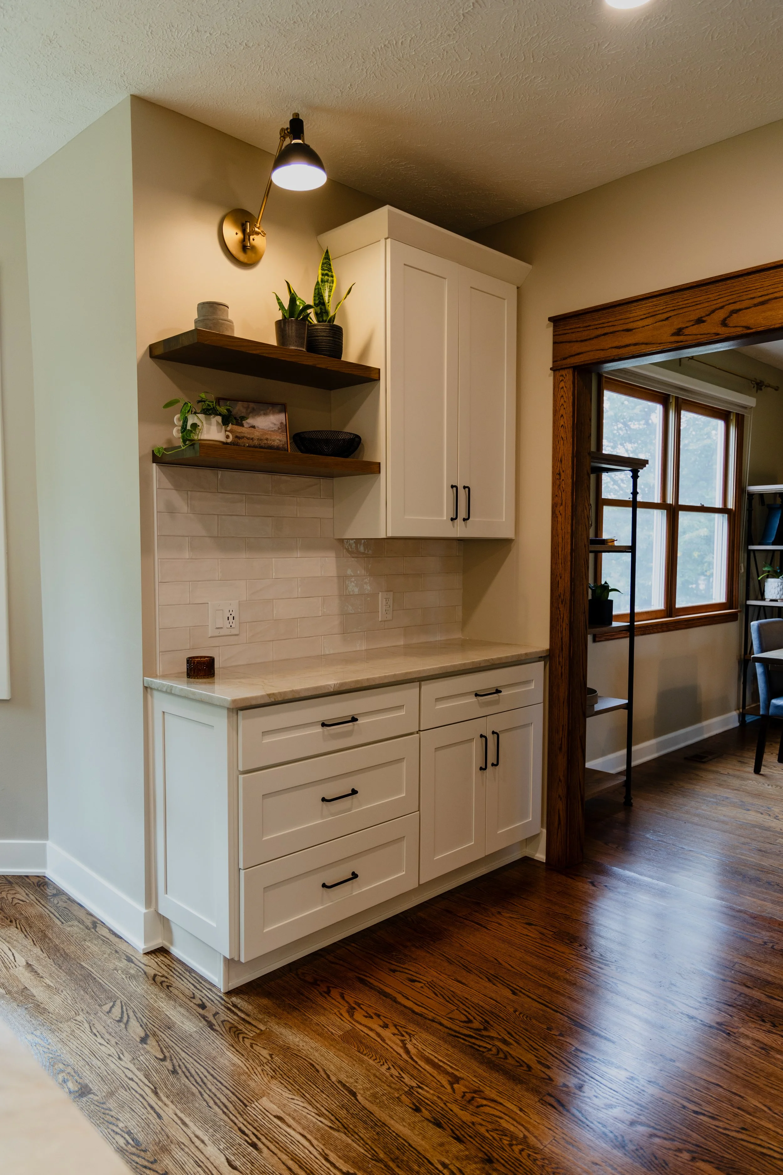 White kitchen cabinet with black handles and marble countertop in a room with wooden flooring, next to a window with a wooden frame and an open doorway.
