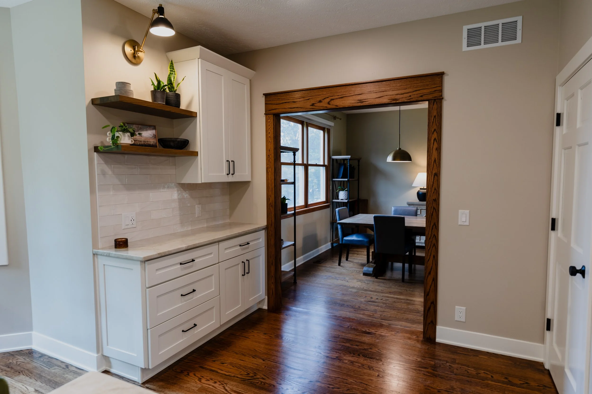 Living room view with white cabinetry, wooden open shelves with plants, and a dining area with a wooden table and chairs, hardwood floors, and window with blinds.