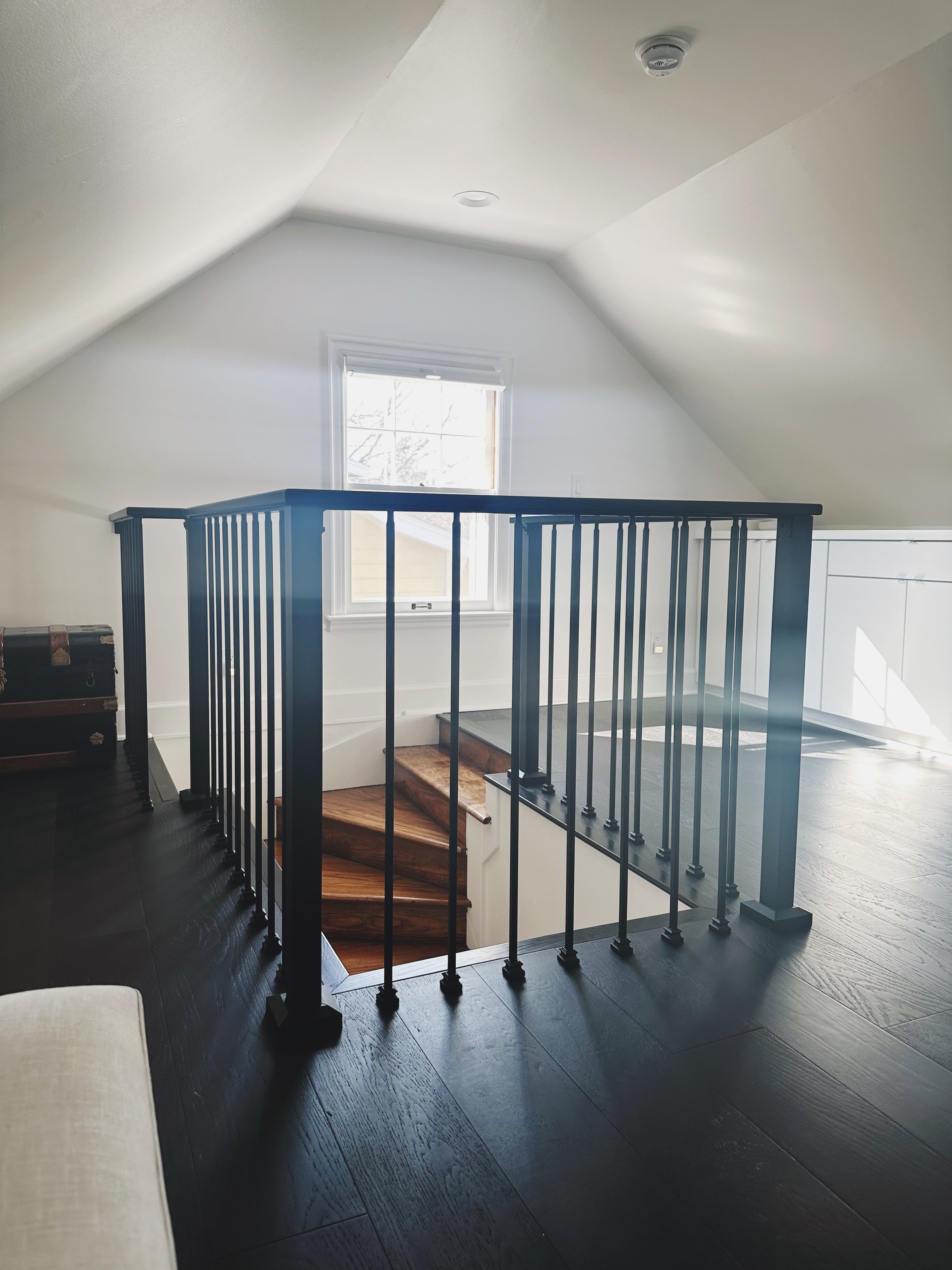 Interior view of a staircase with dark wood steps and black metal railing, leading down to a lower level near a window in a house with white walls and a sloped ceiling.