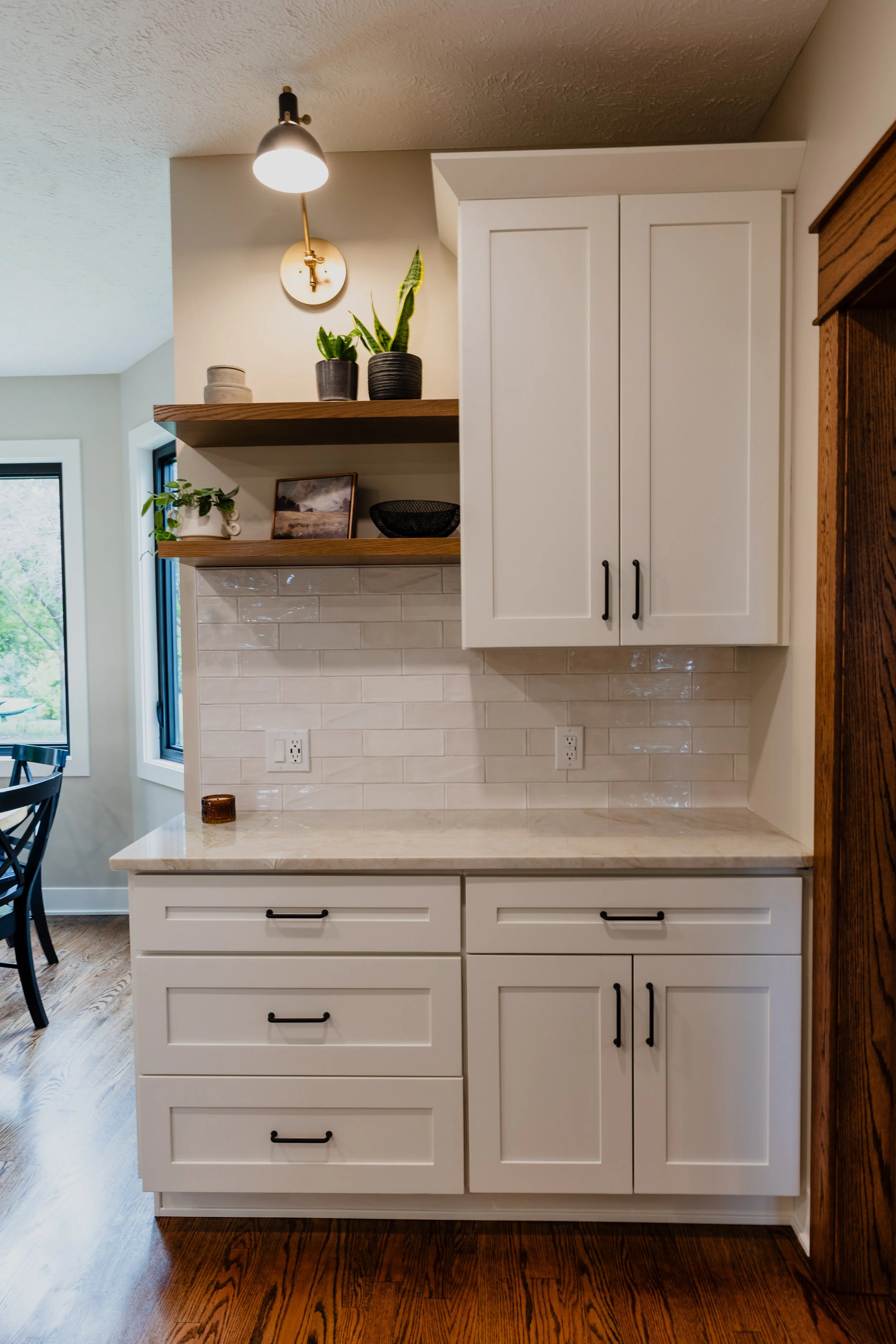 Kitchen cabinetry with upper and lower white cabinets, a marble countertop, two wooden open shelves with plants and decor, white subway tile backsplash, and hardwood flooring.