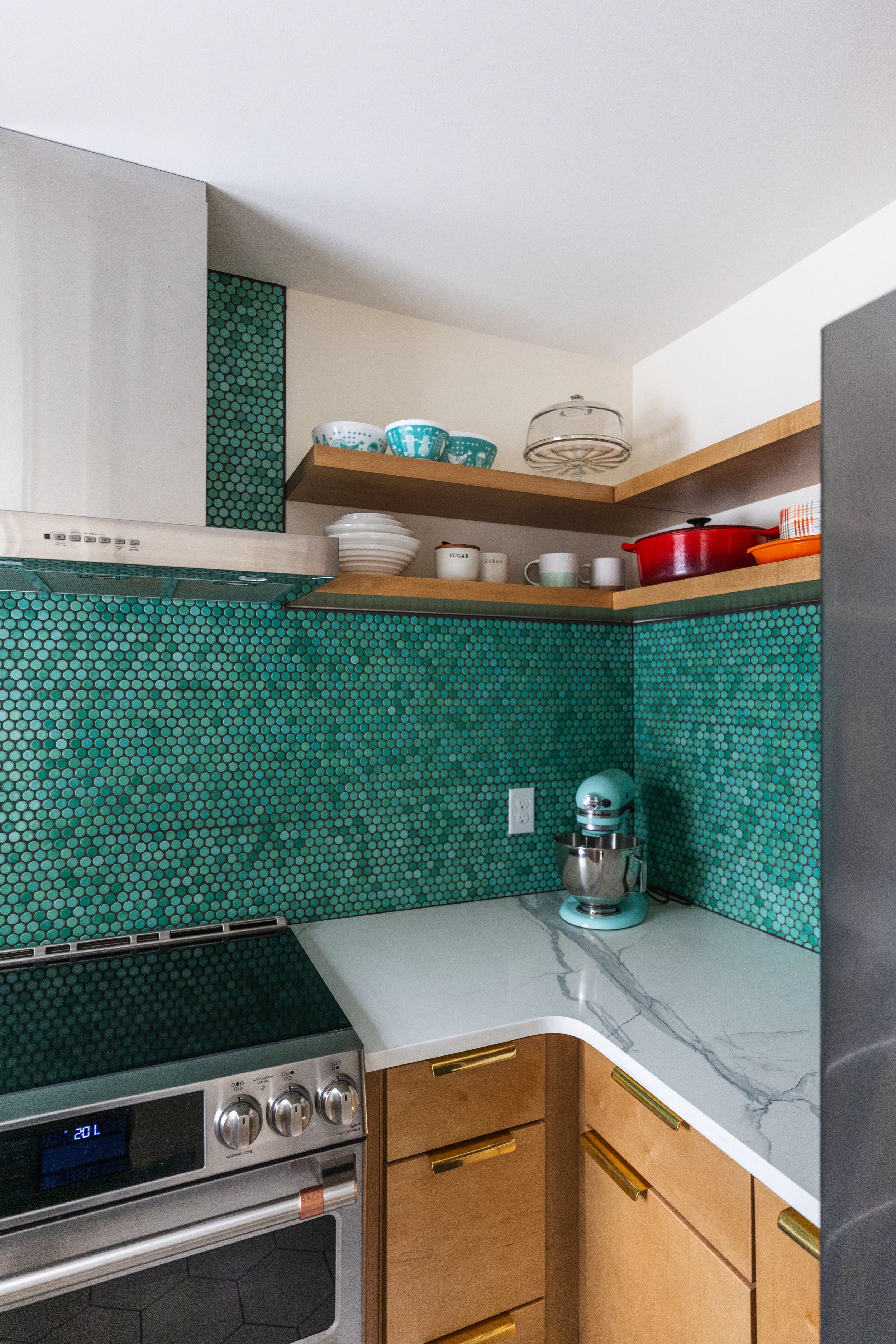 Corner of a modern kitchen with green hexagonal tile backsplash, wooden shelves holding dishes and cookware, and a white marble countertop with teal mixer