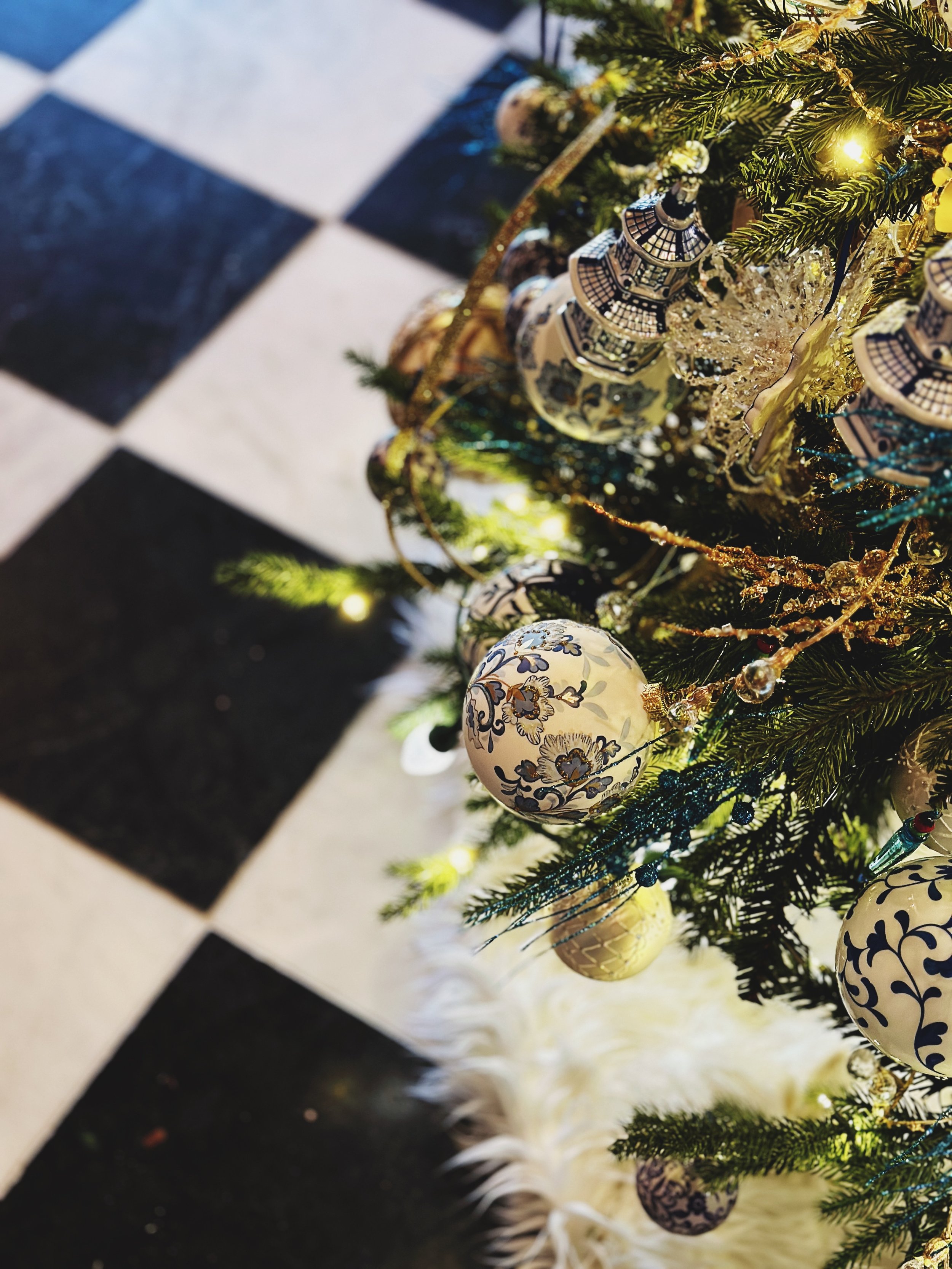 Close-up view of a decorated Christmas tree with ornaments, including patterned baubles and gold accents, on a black and white checkered floor.