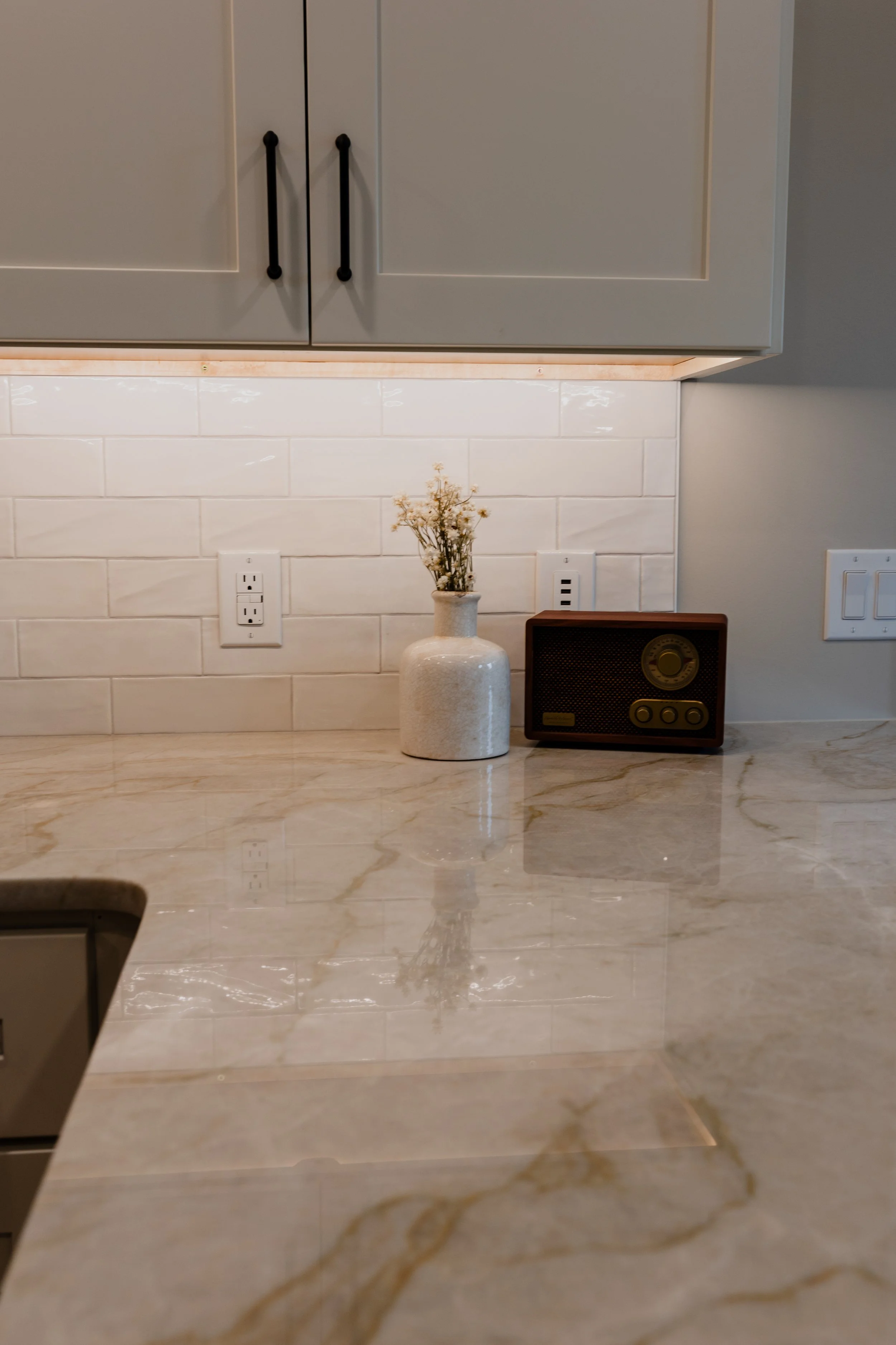 A kitchen countertop with a white vase holding dried flowers and a vintage radio, with white subway tile backsplash and gray cabinetry above. 1935 Kitchen Remodel. Black Matte Hardware. White Cabinets. White Subway Backsplash.