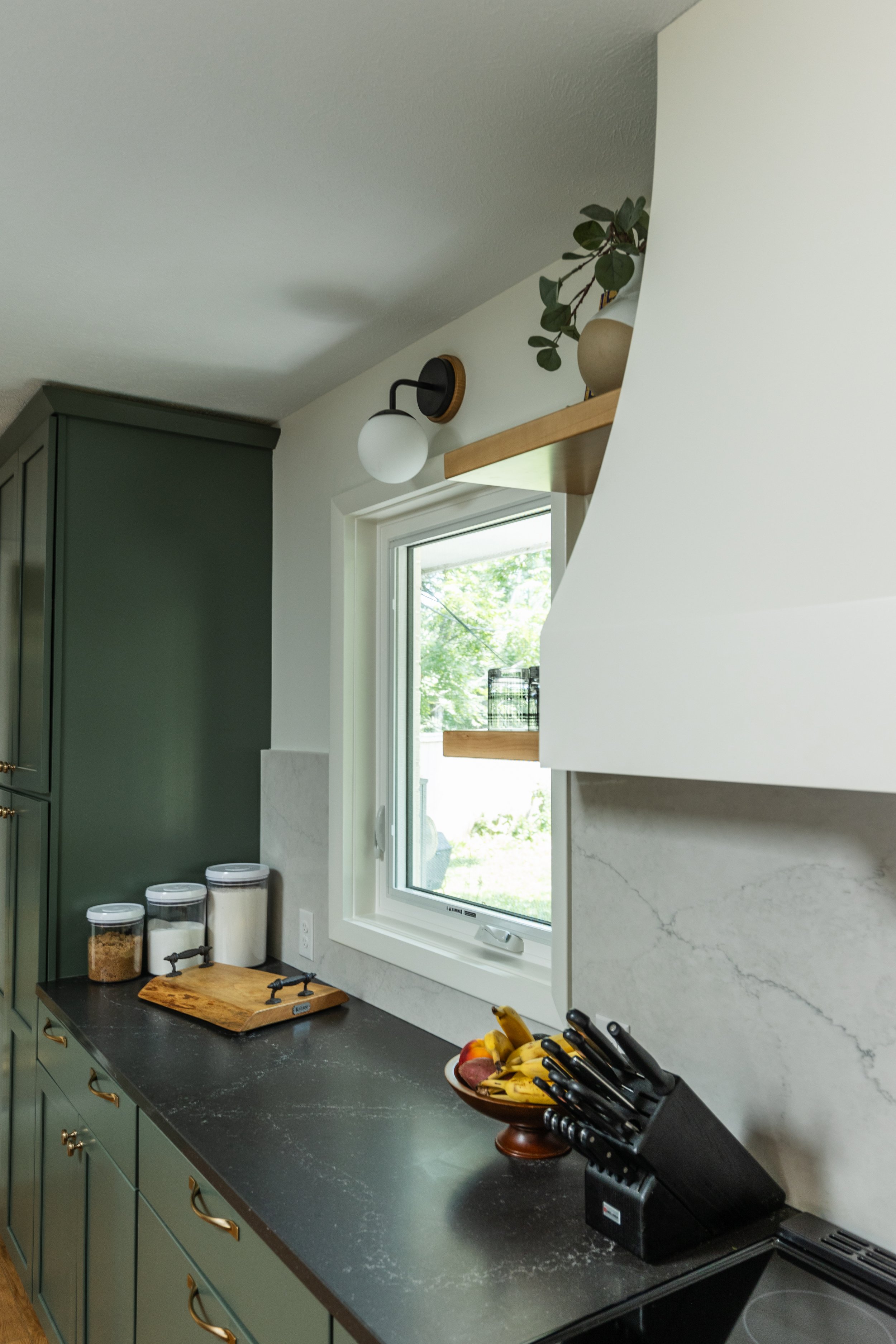 1962 Kitchen Remodel, dark green cabinets, white quartz island, wooden floors, brass hardware, wood cabinets, custom hood, floating shelves, dark countertops, open shelving, large windows.