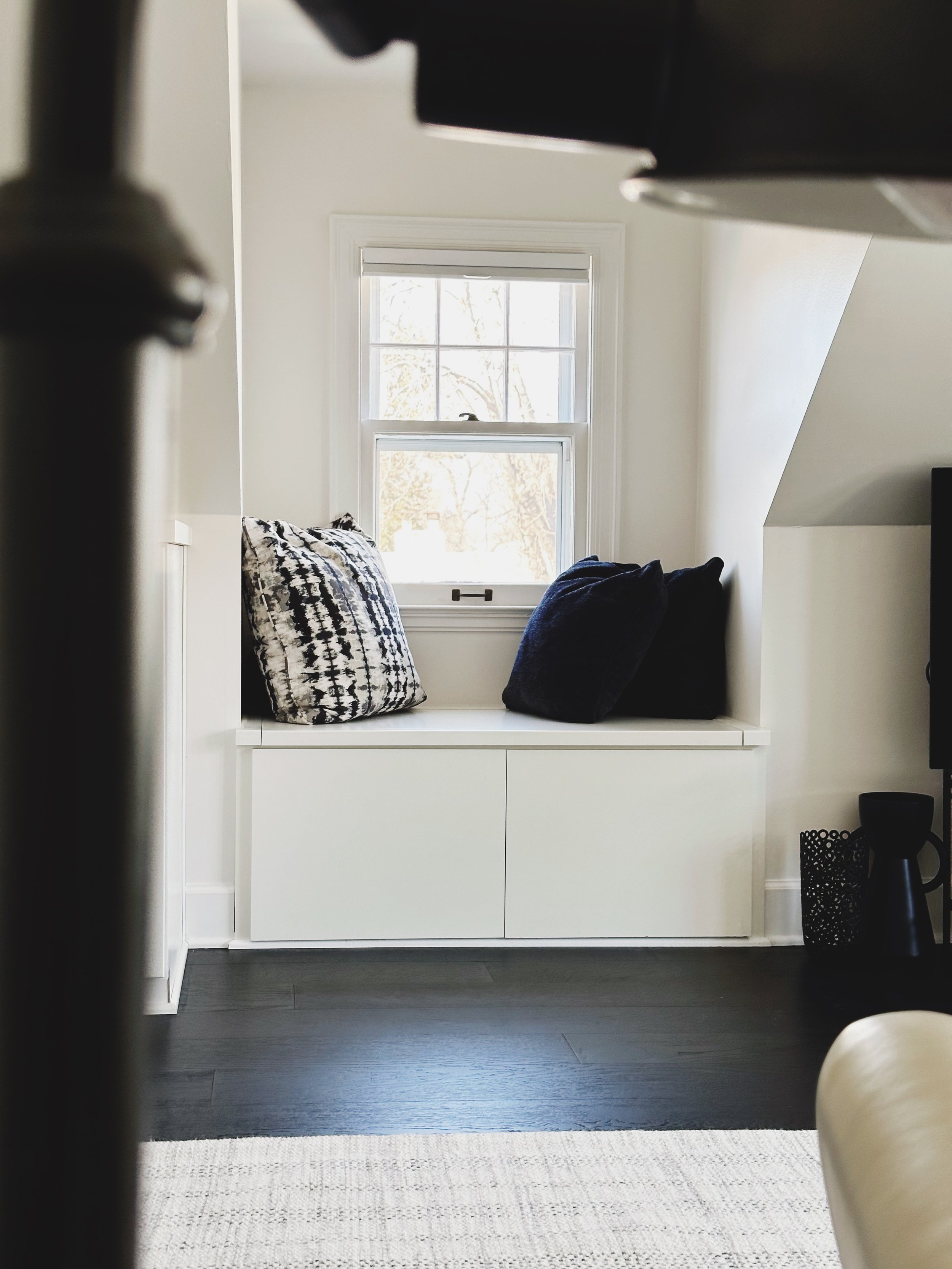 Window seat with three pillows, two dark blue and one patterned black and white, in a bright room with white walls and window, dark hardwood floor, and a beige rug.