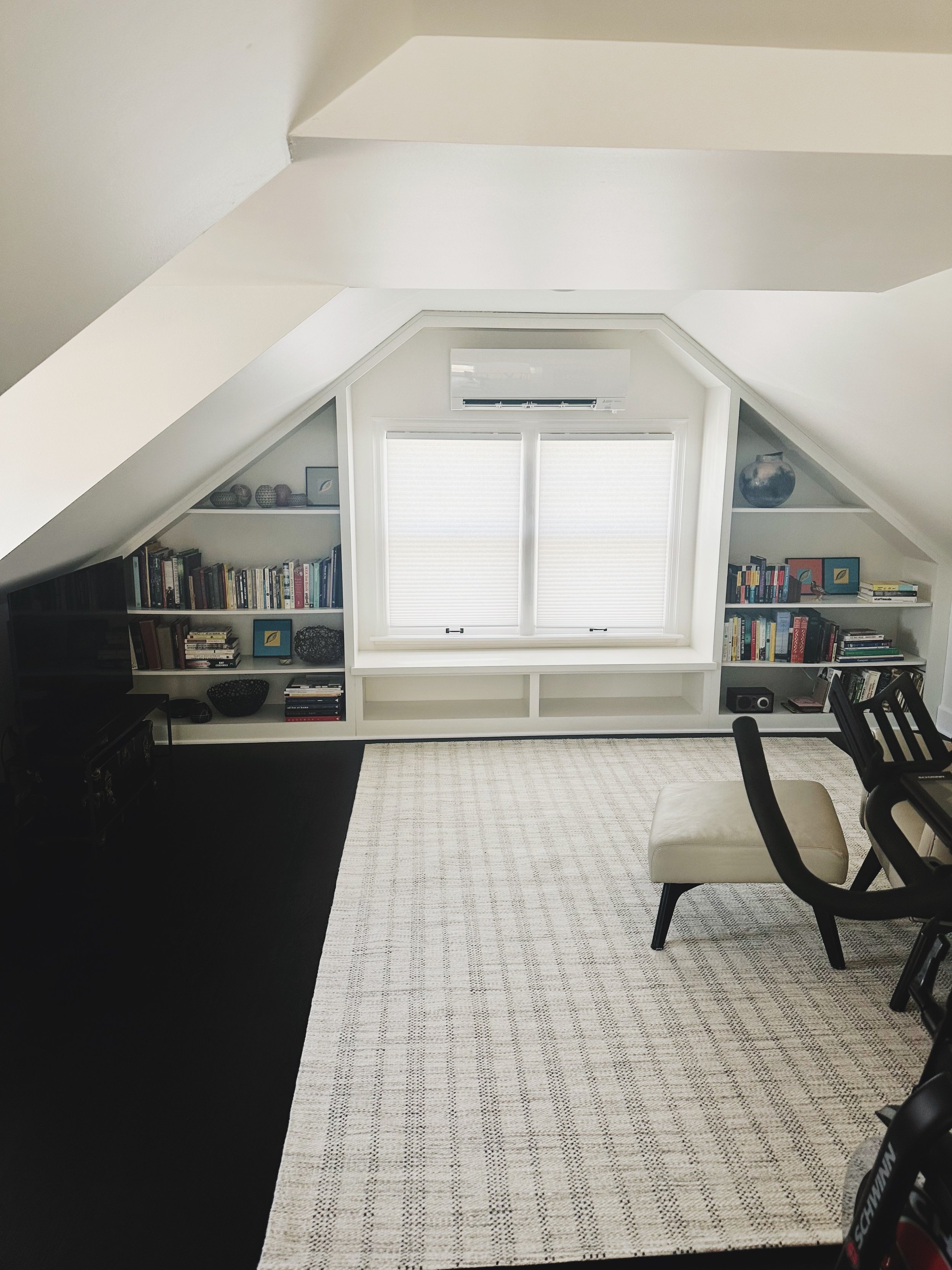 A cozy attic room with built-in white bookshelves on either side of a window, a beige chair, and a black desk in a modern decor setting.