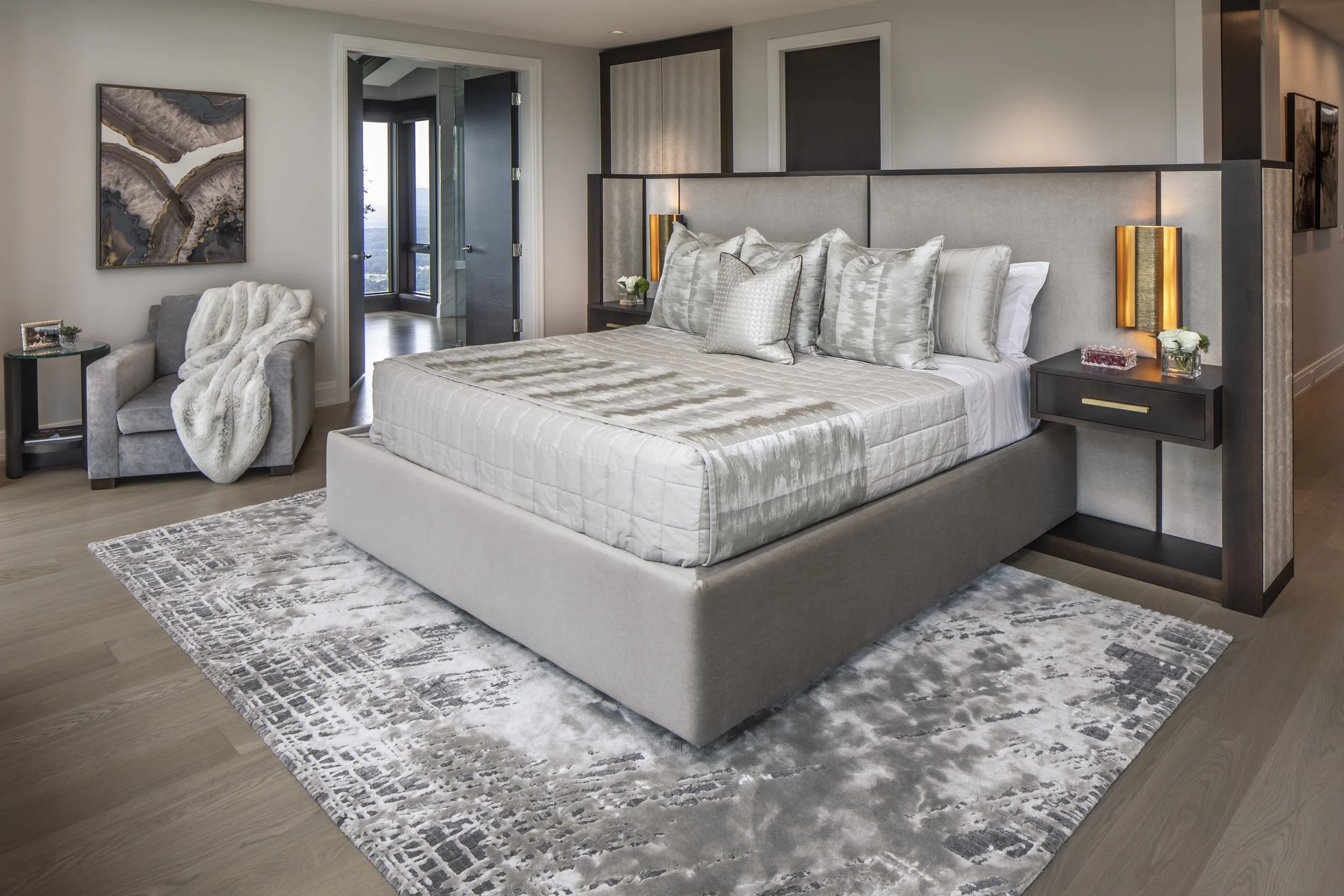 A modern bedroom with a large bed, multiple pillows, side tables with lamps, a patterned area rug, and a sitting chair with a throw blanket, with an open door showing a view outside.