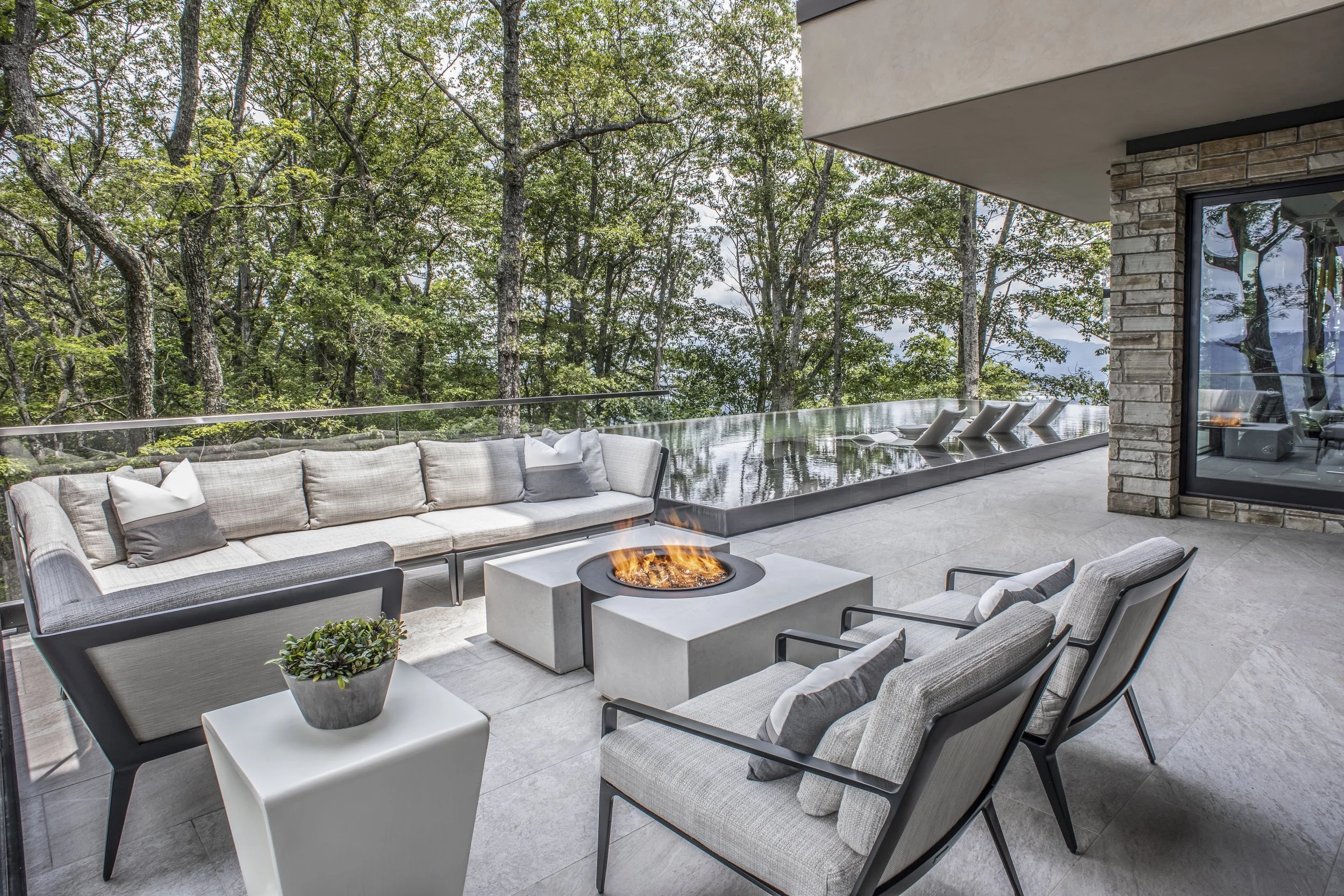 Modern outdoor patio with beige cushioned seating, lounge chairs, a fire pit, and a glass railing overlooking a wooded area.