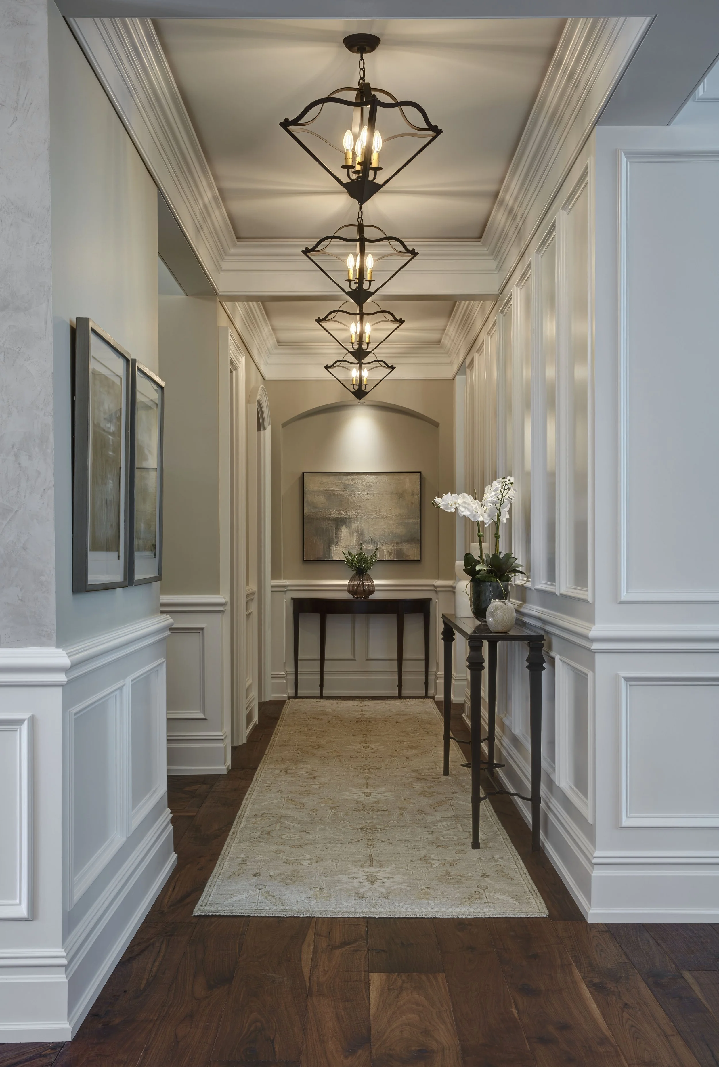 Elegant interior hallway with white paneled walls, hardwood flooring, and three chandelier-style light fixtures hanging from ceiling. There is a beige rug on the floor, a console table with a plant in a vase, and artwork on the wall.