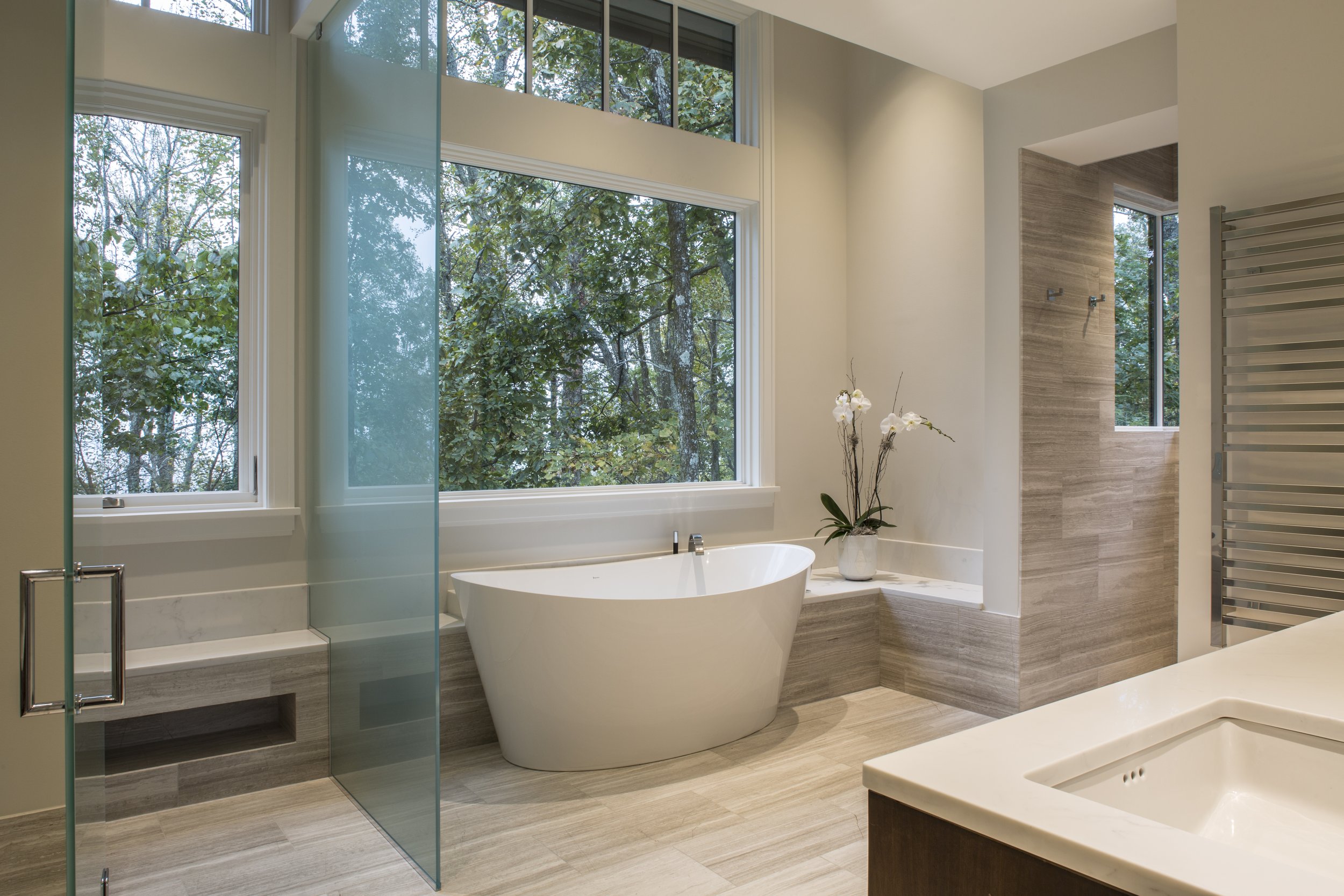 Modern bathroom with a freestanding bathtub placed by large windows overlooking a wooded outdoor scene. There is a vase with orchids on the ledge near the tub. A walk-in shower with glass doors and wood-textured tiles is visible, along with a heated 