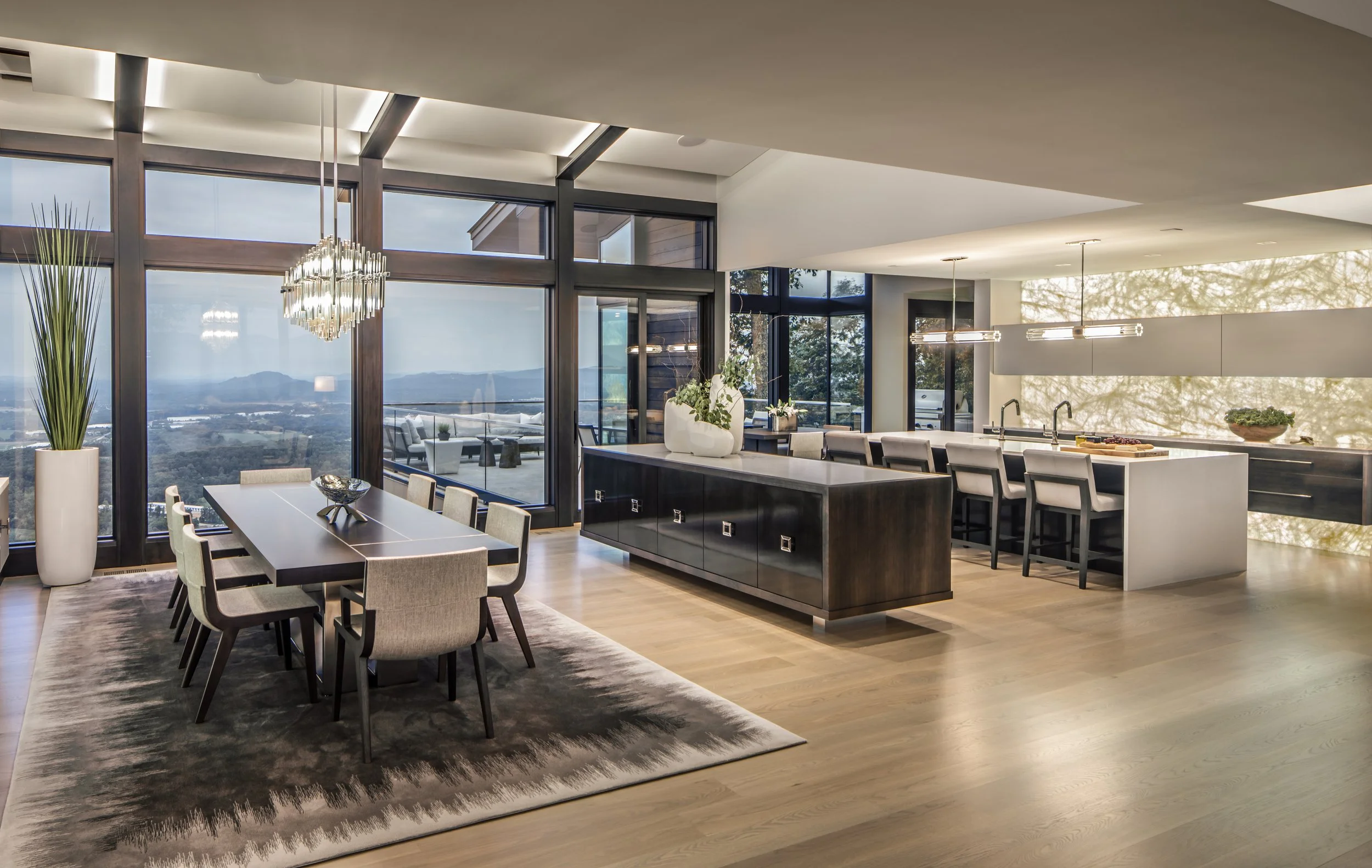 Open-concept dining and kitchen area with large windows, modern chandelier, central island with seating, and outdoor patio view