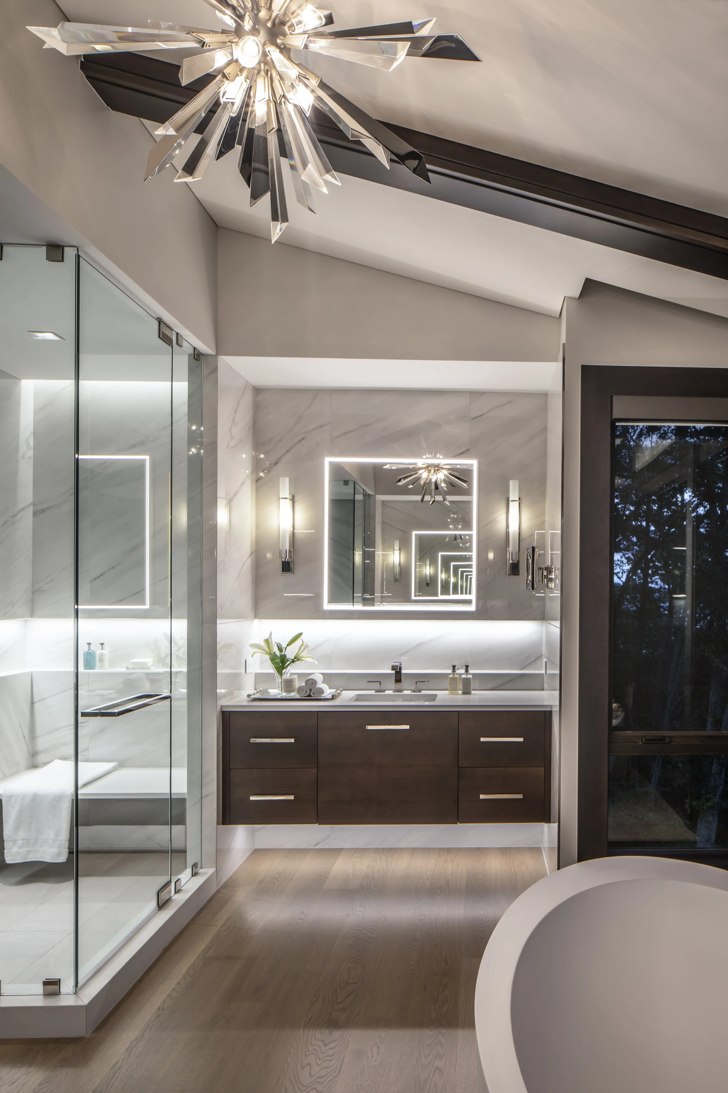 A modern bathroom with a wooden vanity, large mirror with LED lighting, glass-enclosed shower, bathtub, and contemporary chandelier in a well-lit space.
