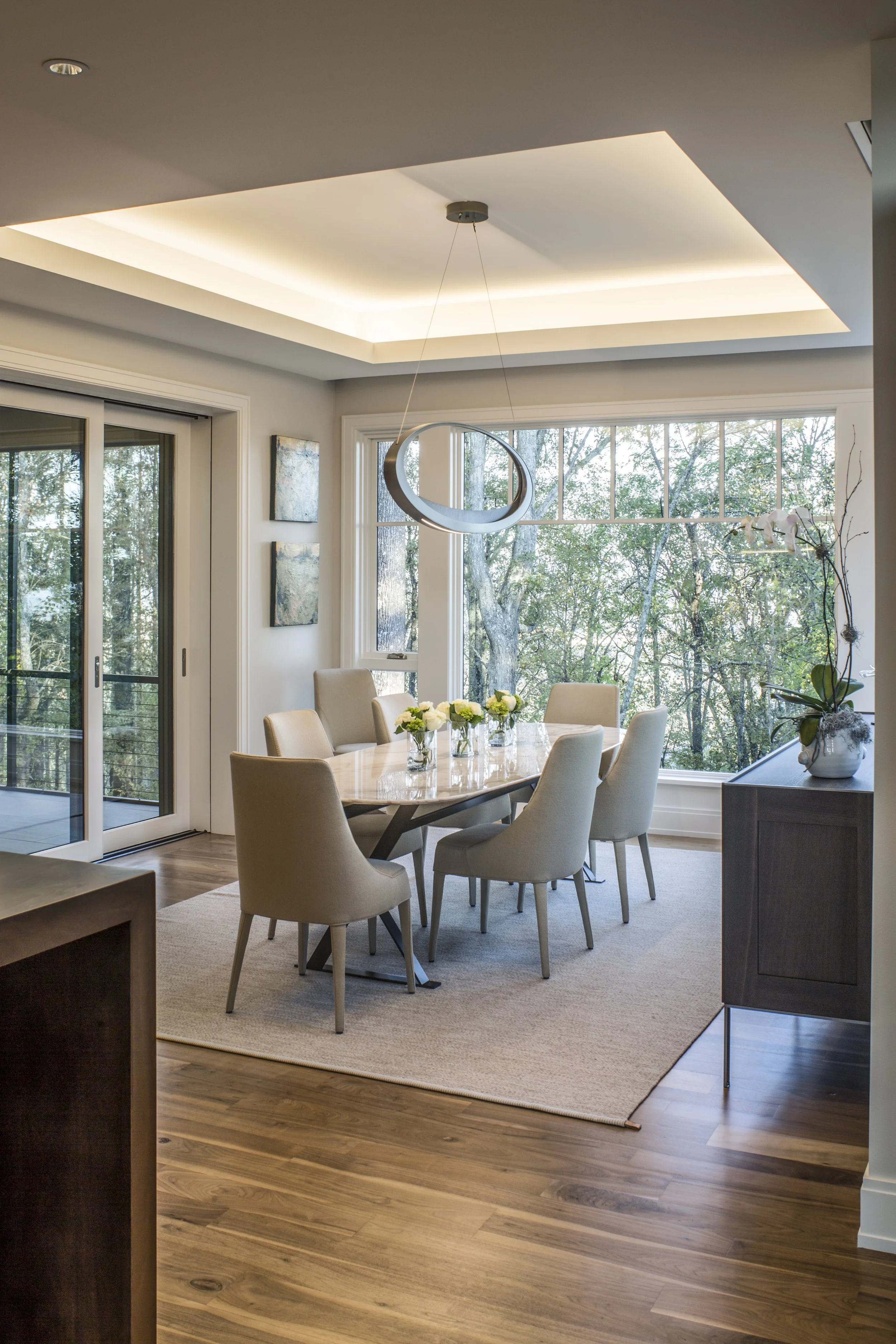 Modern dining room with a wooden table, beige chairs, large window with trees outside, hanging light fixture, and artwork on the wall.