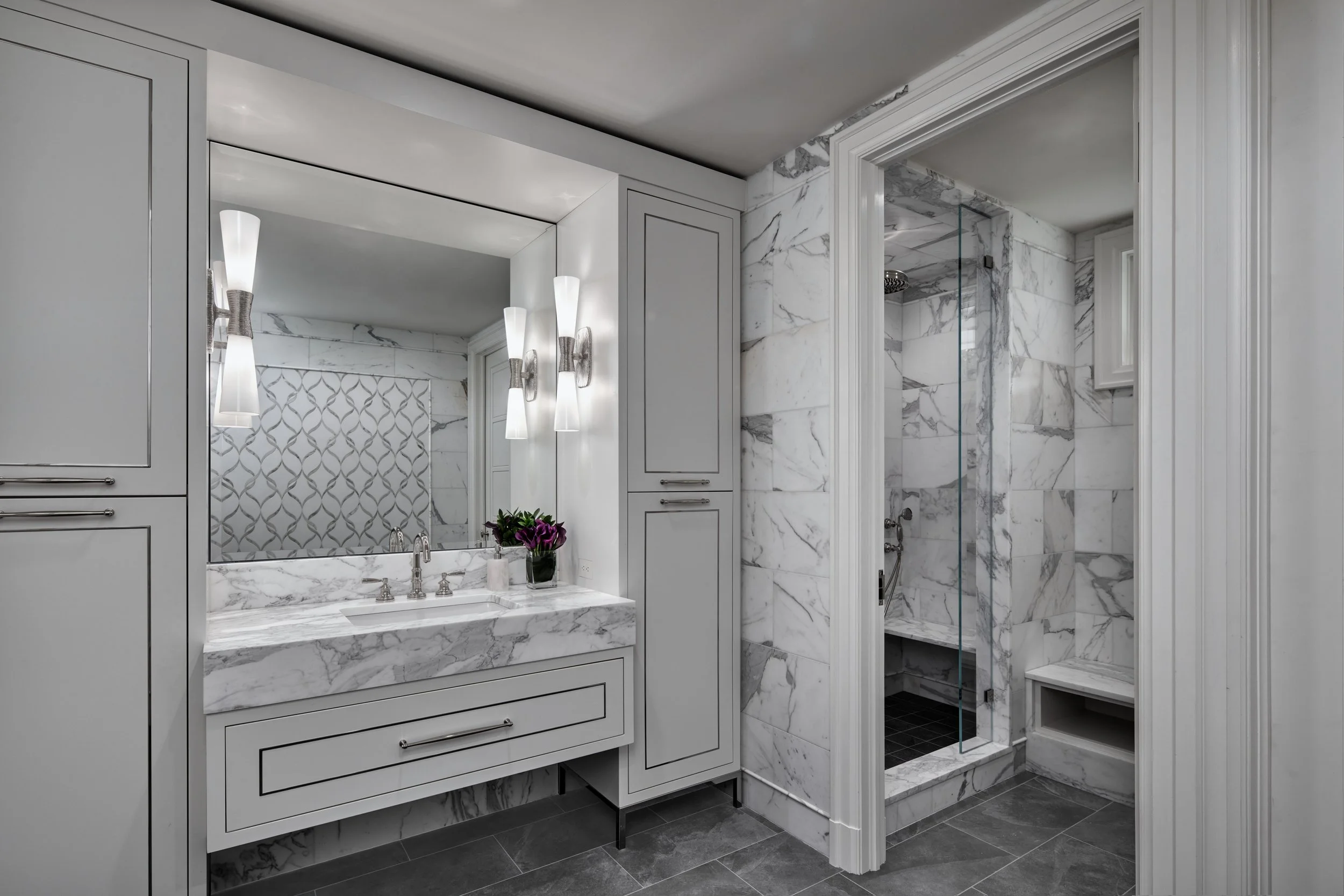 A modern bathroom with marble walls and floor, featuring a large mirror, a white vanity with a marble top, and a potted plant. The shower area is enclosed with clear glass and has a marble interior and black tiles on the floor.