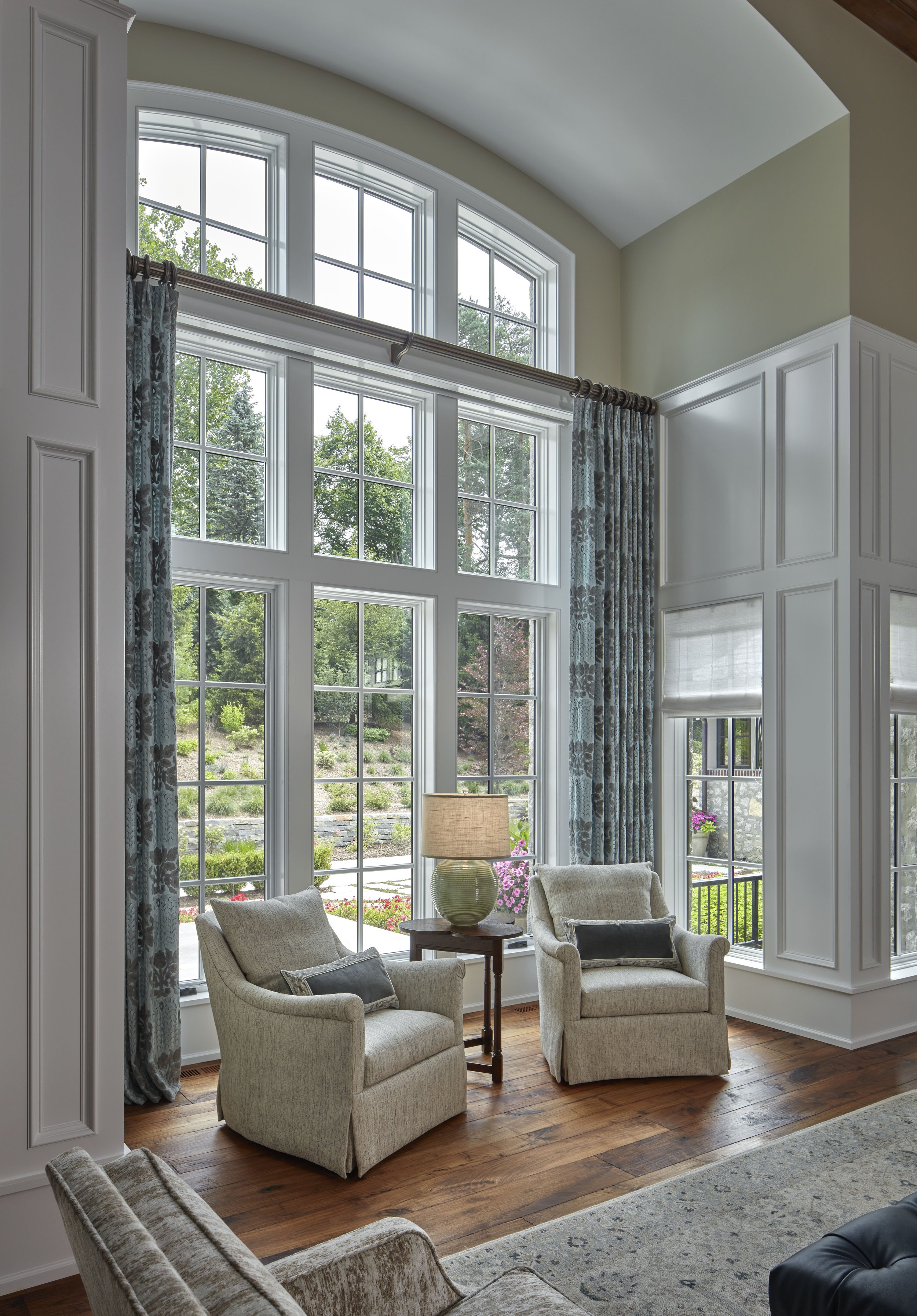 Living room with large bay window, beige armchairs, wooden floor, and patterned curtains.