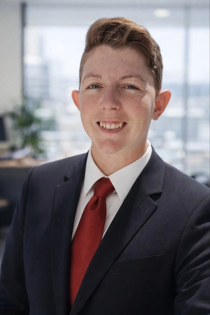 A young man in a suit and red tie smiling in an office setting with windows and blurred background.