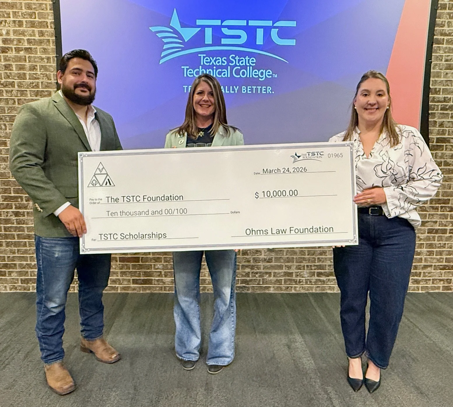 Three women and one man standing indoors holding a large check made out to TSTC Foundation for $10,000. Behind them is a screen displaying the TSTC logo and text. The brick wall is visible in the background.