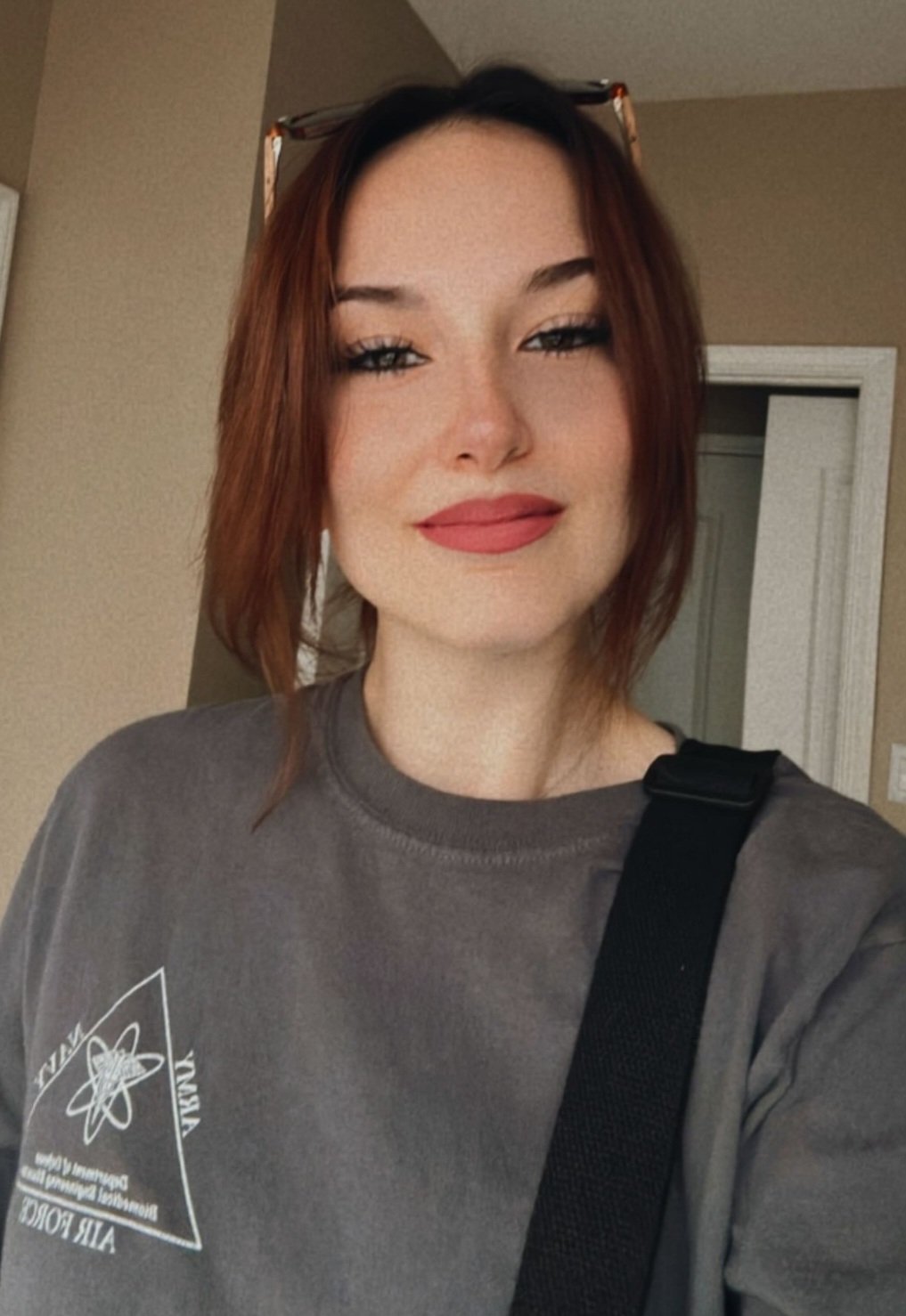 A woman with red hair, wearing a gray t-shirt and sunglasses on her head, smiling at the camera inside a home.