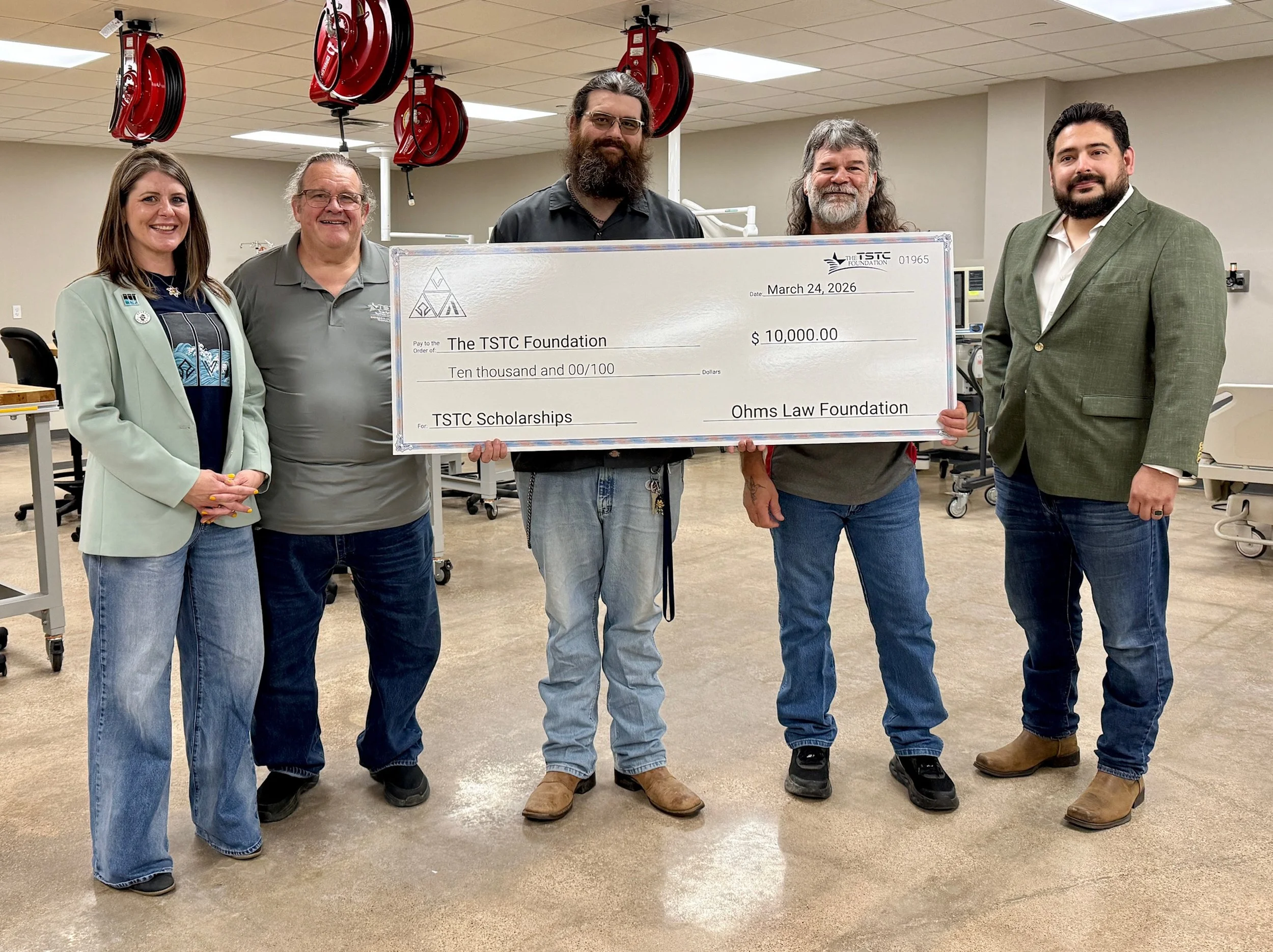 Five people standing indoors holding a large check for $10,000 made out to The TSTC Foundation for scholarships, with red hoses hanging from the ceiling in the background.