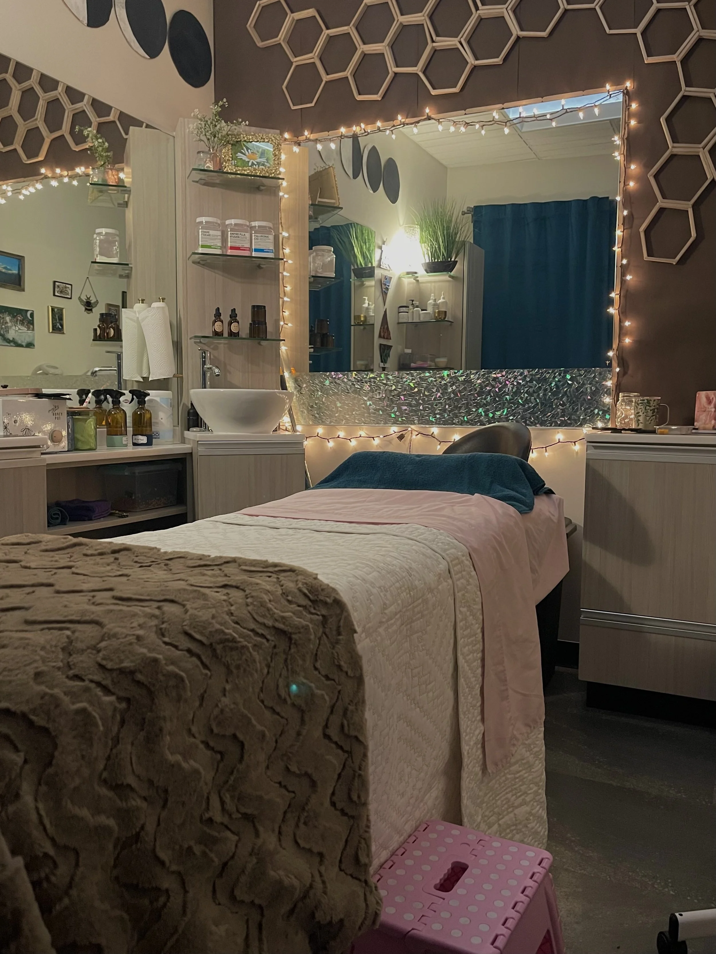 A massage or spa room with a massage bed covered in a pink and white quilt, with a brown textured blanket and a teal towel on top. Behind the bed is a large mirror with string lights, and the room features hexagon wall decorations and shelves with various bottles and plants.