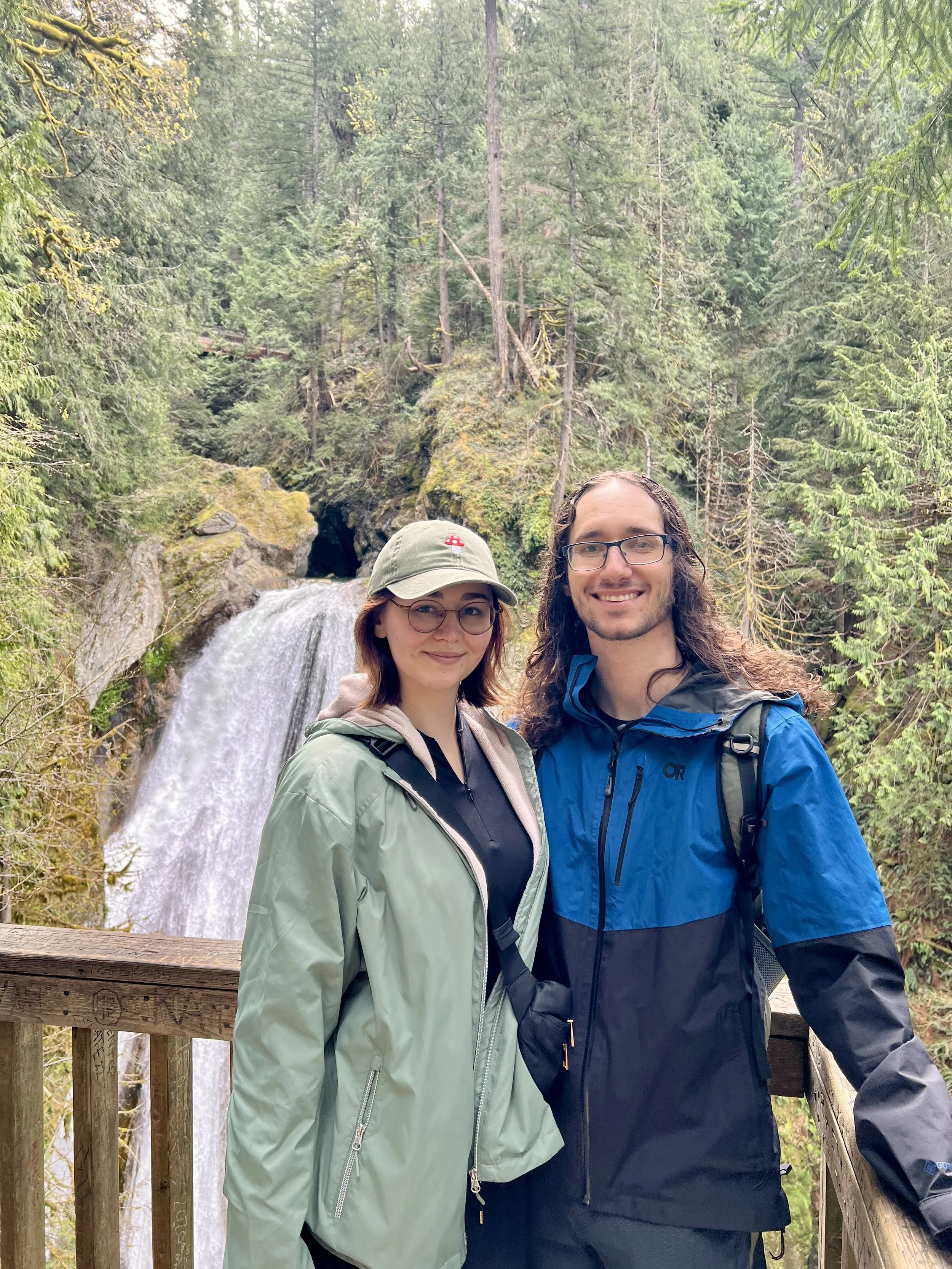 Two people standing on a wooden railing in front of a waterfall surrounded by dense forest, smiling at the camera.
