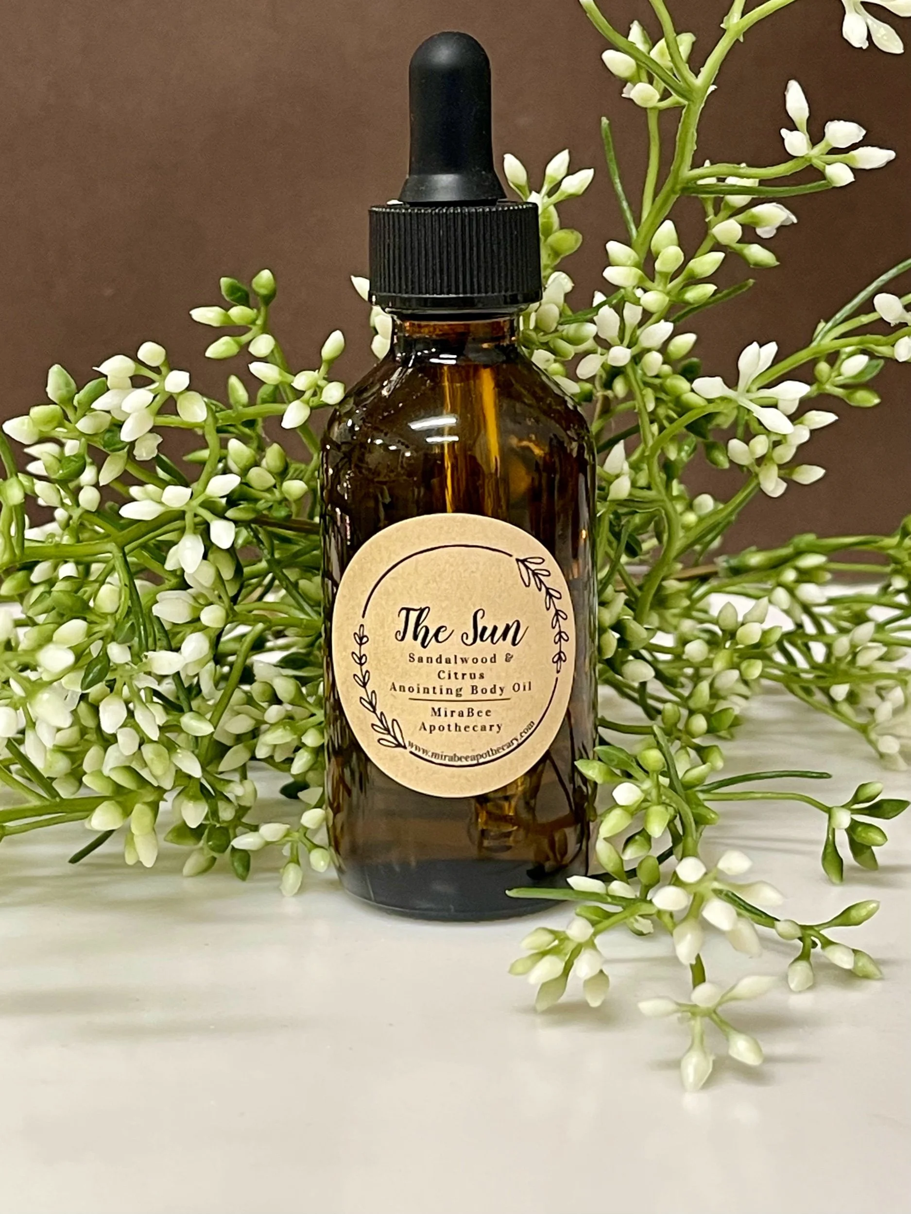 A bottle of The Sun Sandlewood & Citrus Anointing Body Oil placed among white flowering branches on a white surface with a brown background.