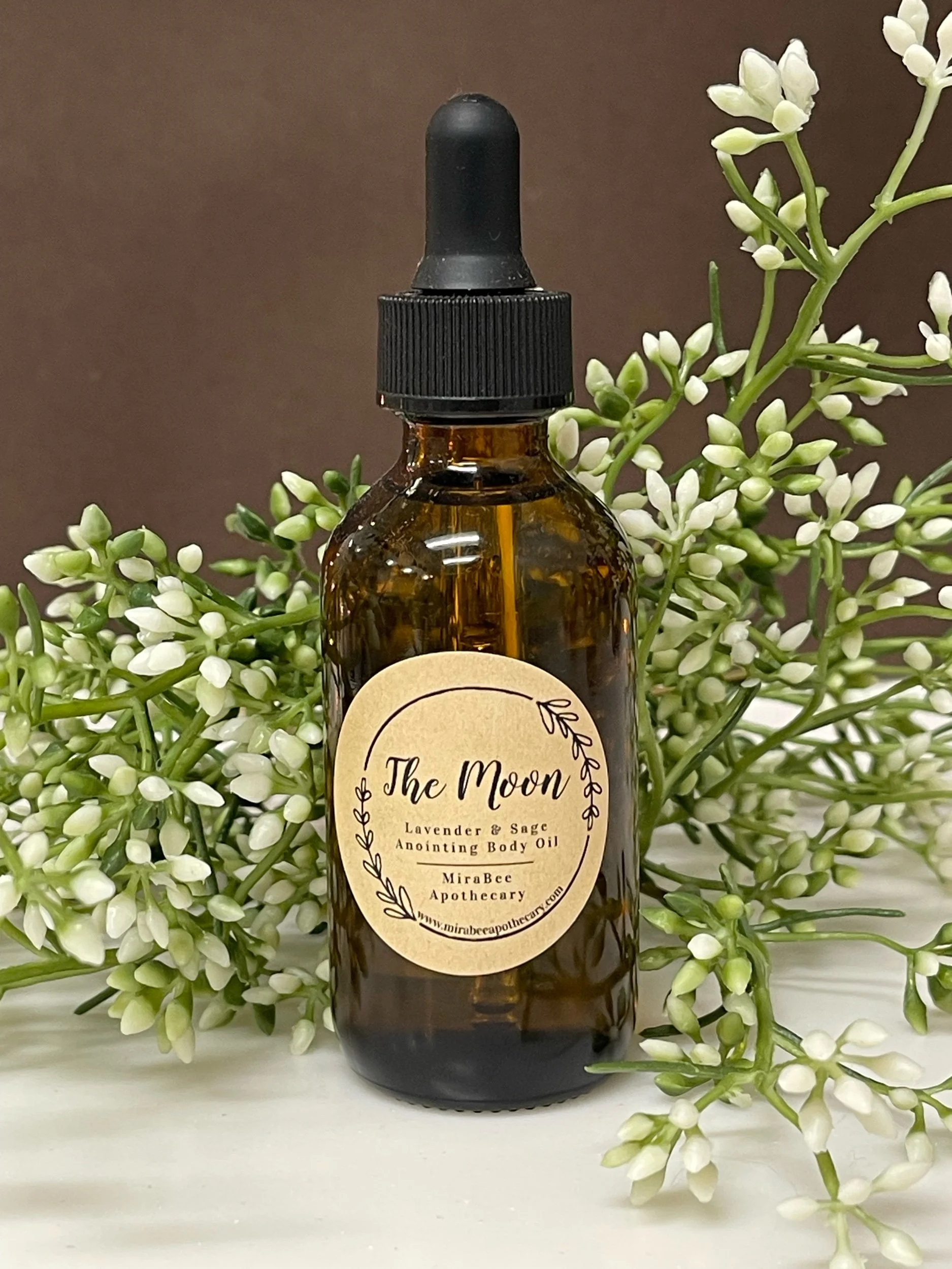 A brown glass bottle of essential oil with a black dropper cap, labeled "The Moon Lavender & Sage Anointing Body Oil, MiraBee Apothecary" sitting among white and green flowers, with a plain neutral background.