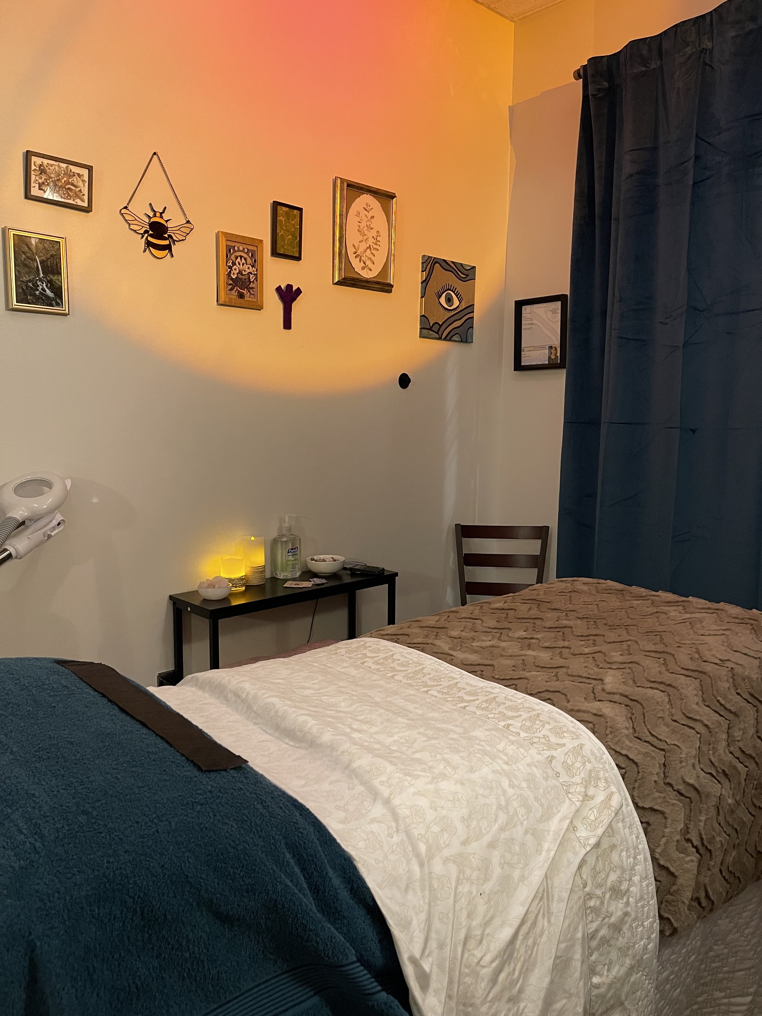 A room with two beds, a black nightstand with candles and bottles, framed art on the wall, a wooden chair, and dark curtains.