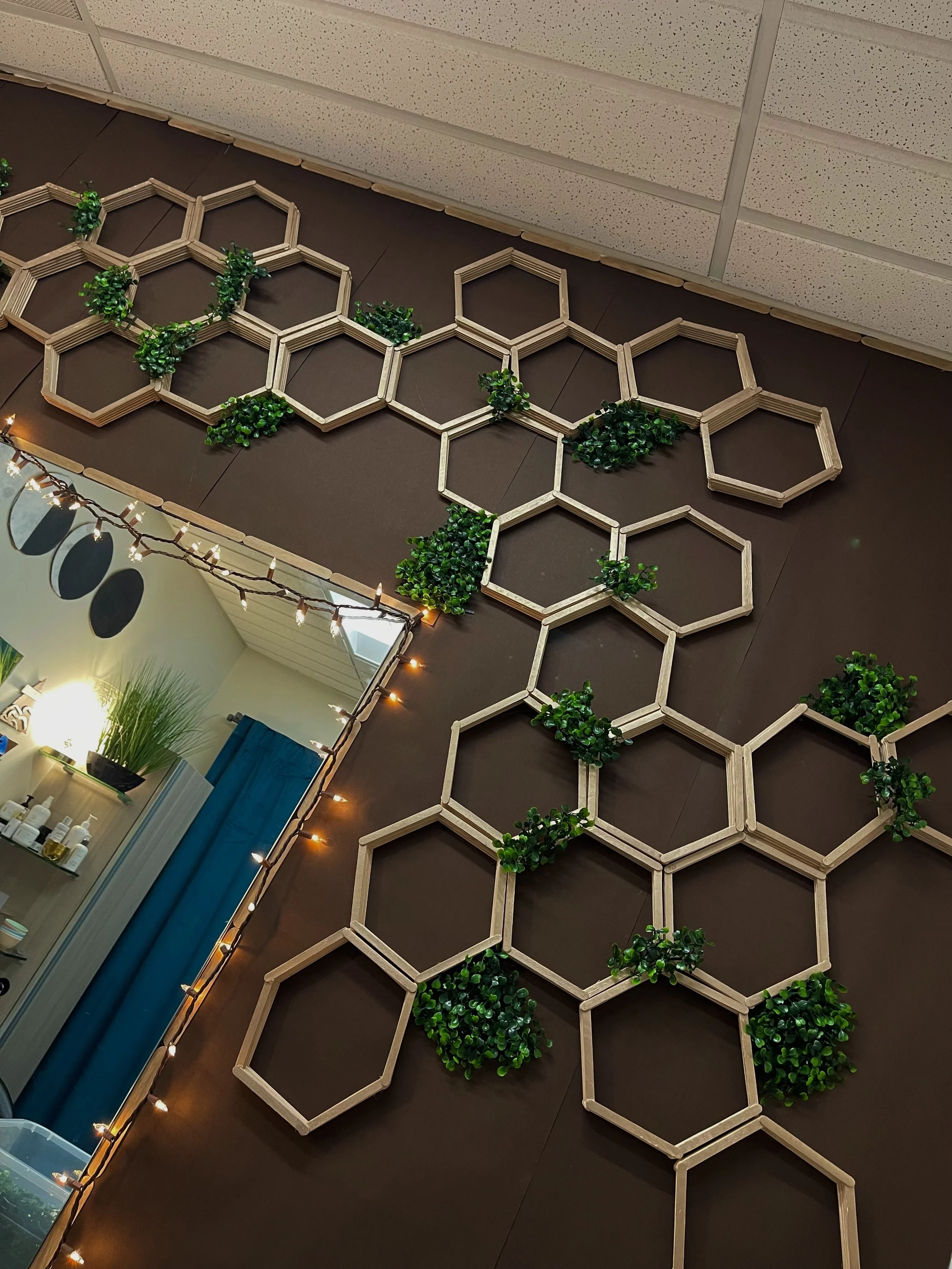 Decorative wall with interconnected wooden hexagons and green plants, illuminated with string lights, in an indoor space.